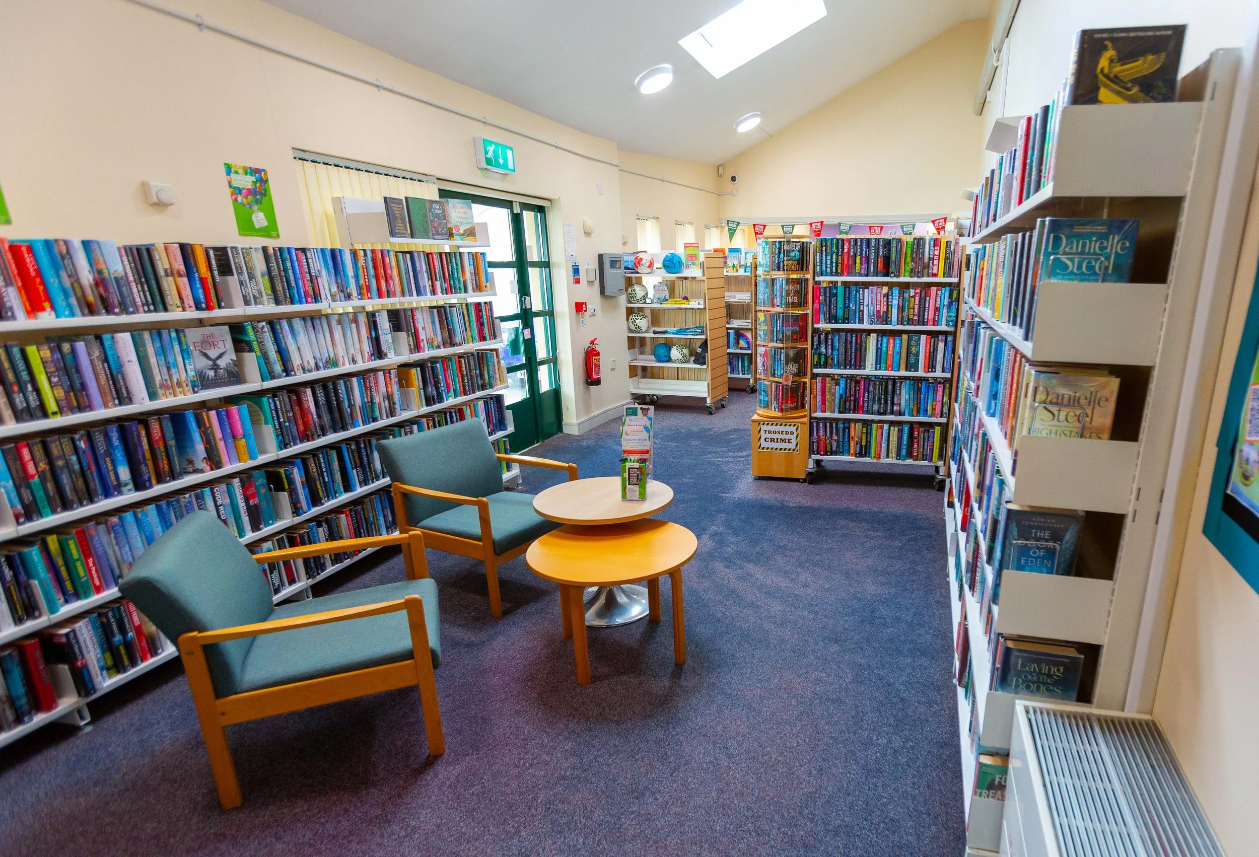 Interior of a bookstore or library with bookshelves filled with books, seating area with two chairs and a round table, illuminated by skylights and ceiling lights.