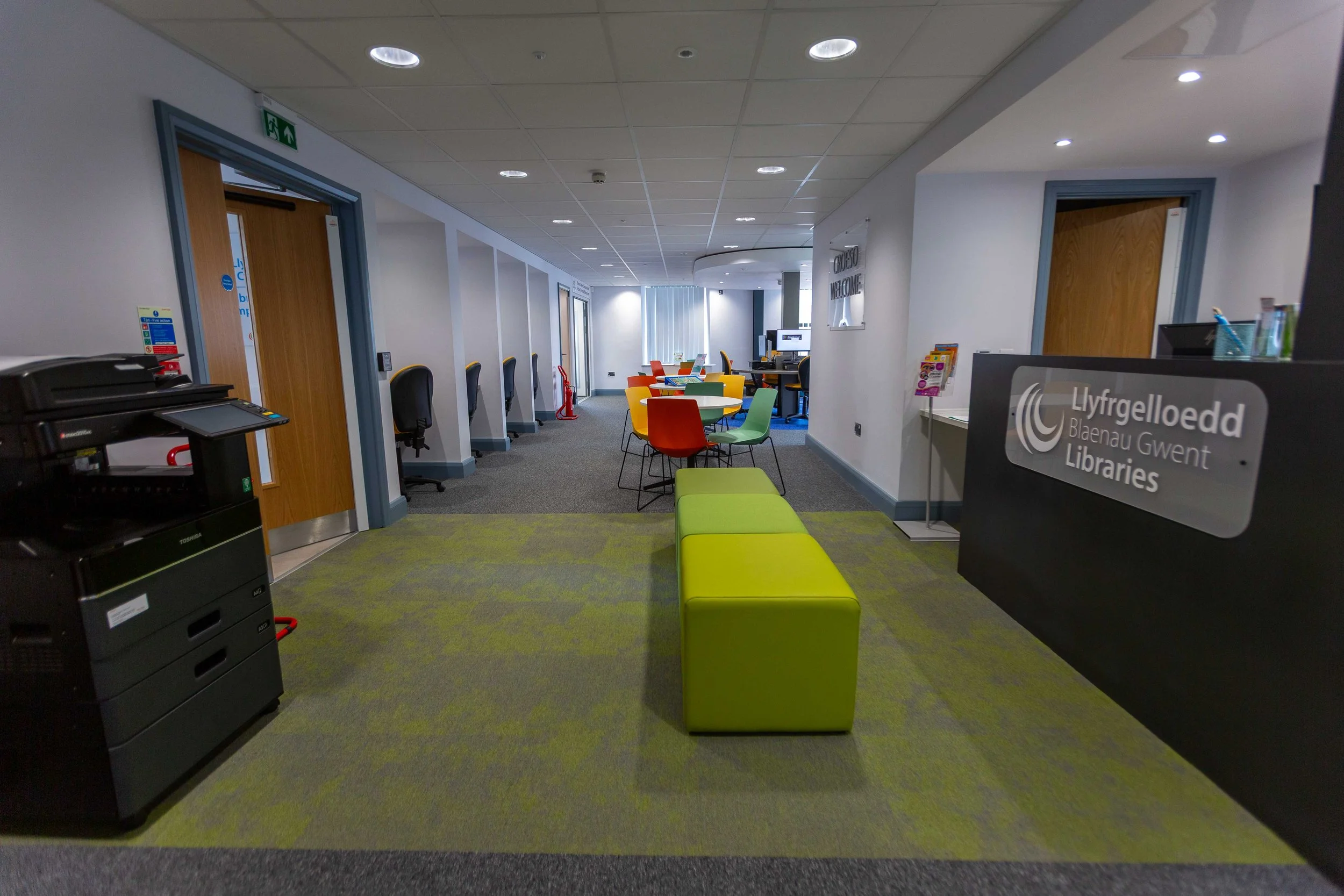 Interior view of Bleanau Gwent Libraries, showing a reception desk on the right, a waiting area with colorful chairs, and computer workstations along the left wall. The space is well-lit with modern decor.