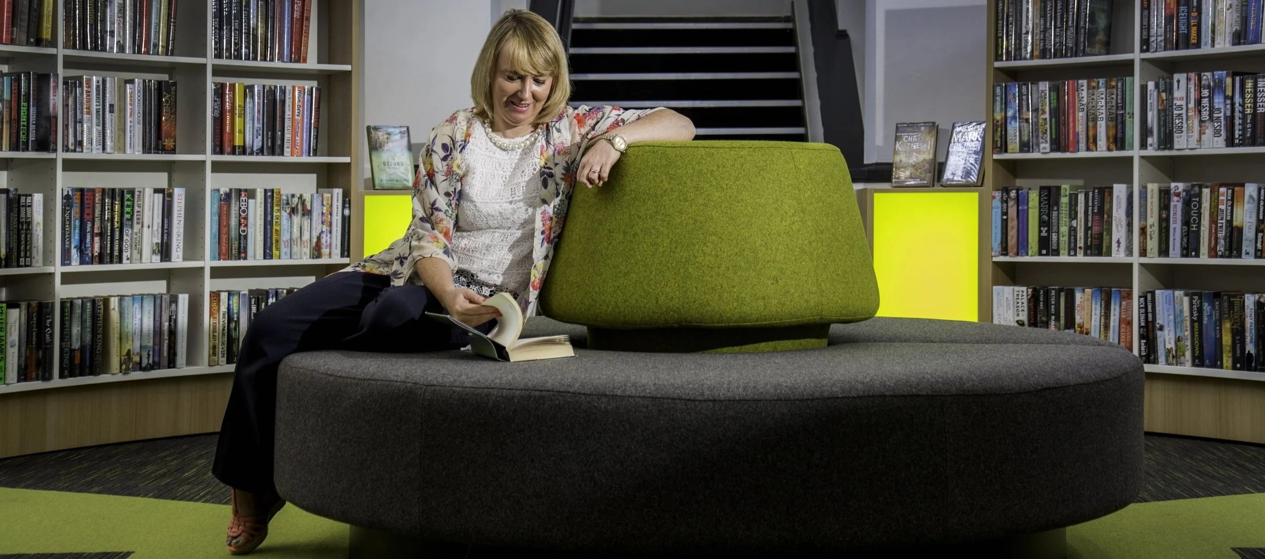 A woman with blonde hair sitting on a round gray bench in a library, looking at a book and smiling.