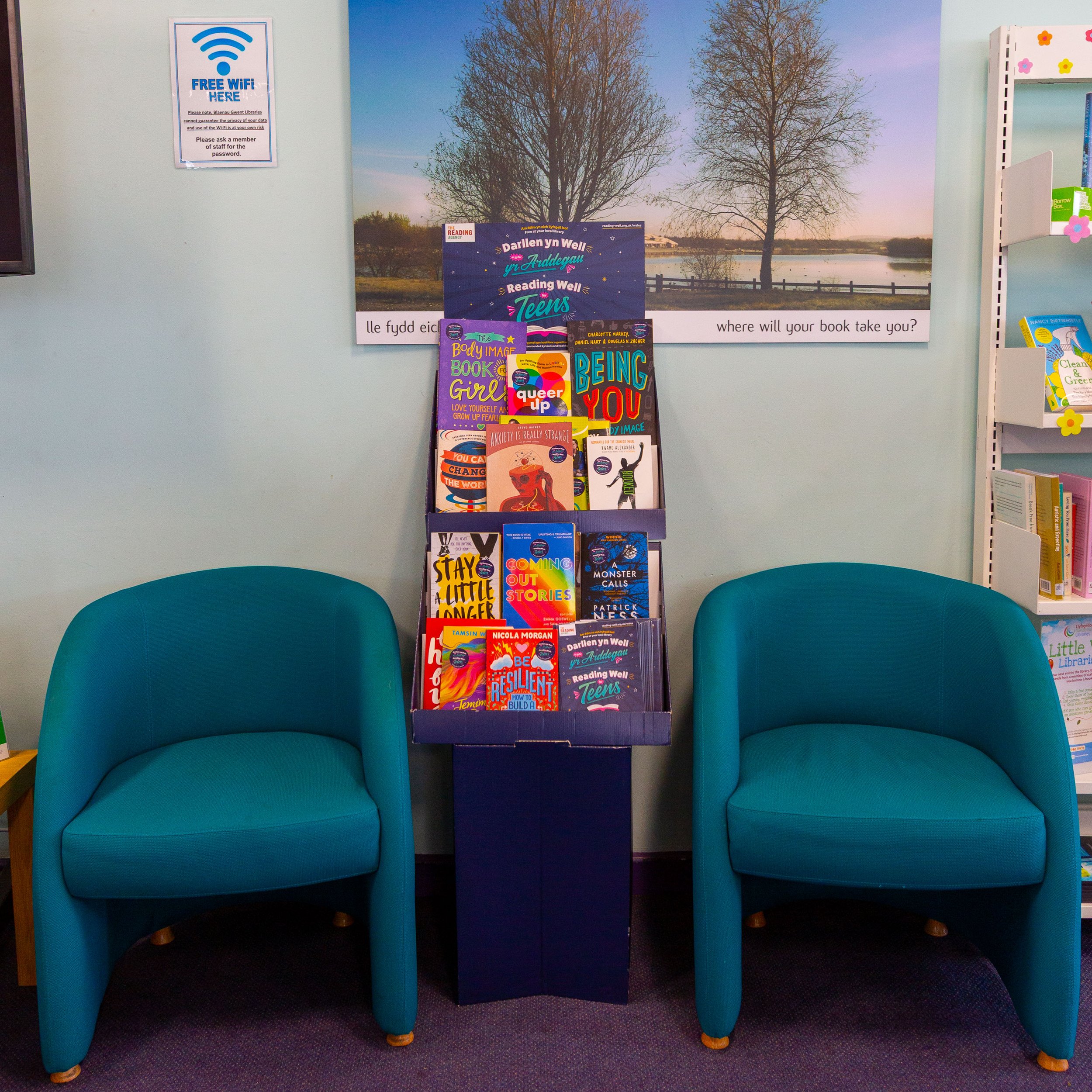 Bookshelf with children's books between two turquoise chairs in a library. Wall decor includes a landscape picture of trees and water, and a sign offering free Wi-Fi.