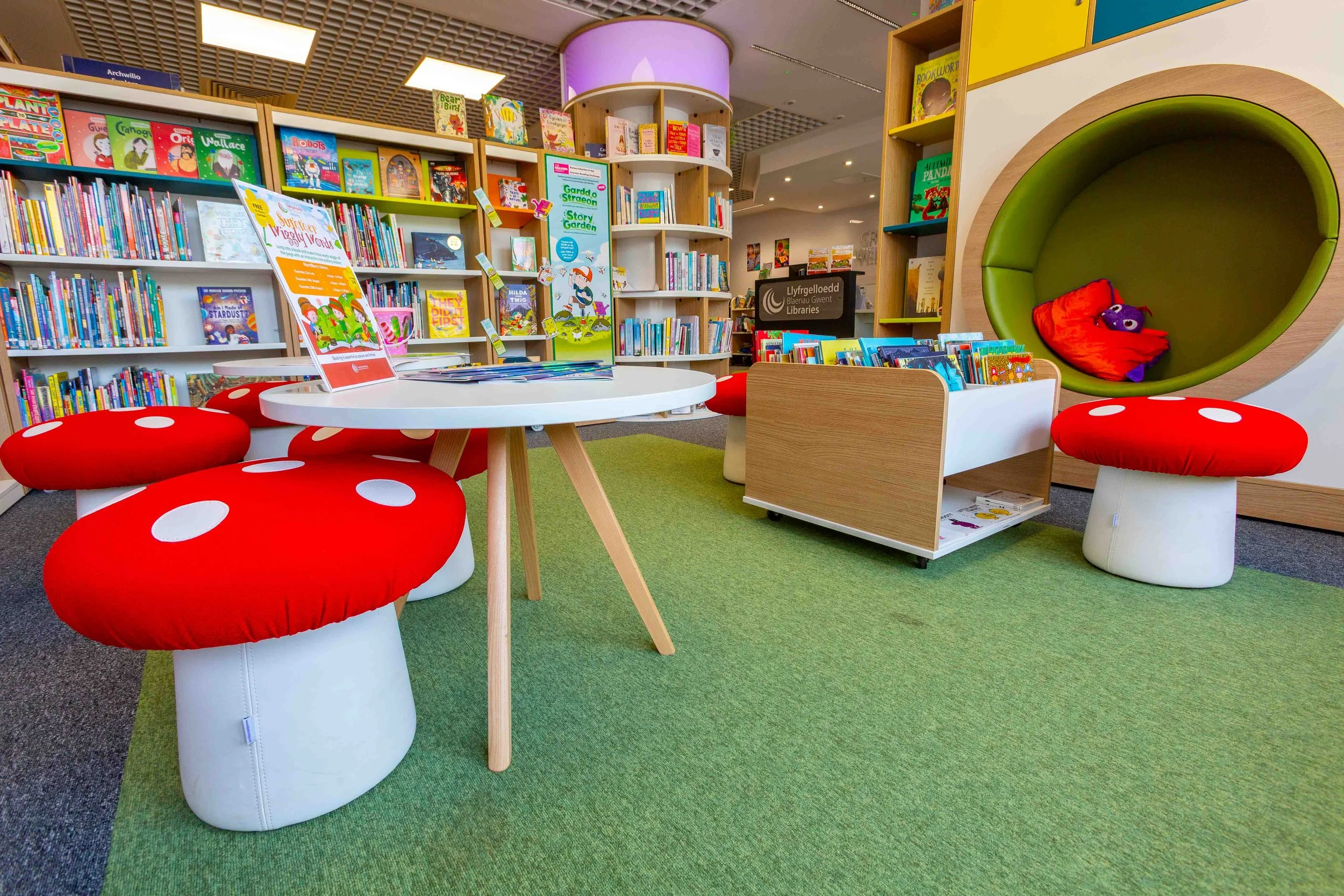 Colorful children's section of a library with bookshelves filled with children's books, red mushroom-shaped seats, a round white table with children's books and a poster, and a cozy green circular reading nook with a purple plush toy and red blanket.