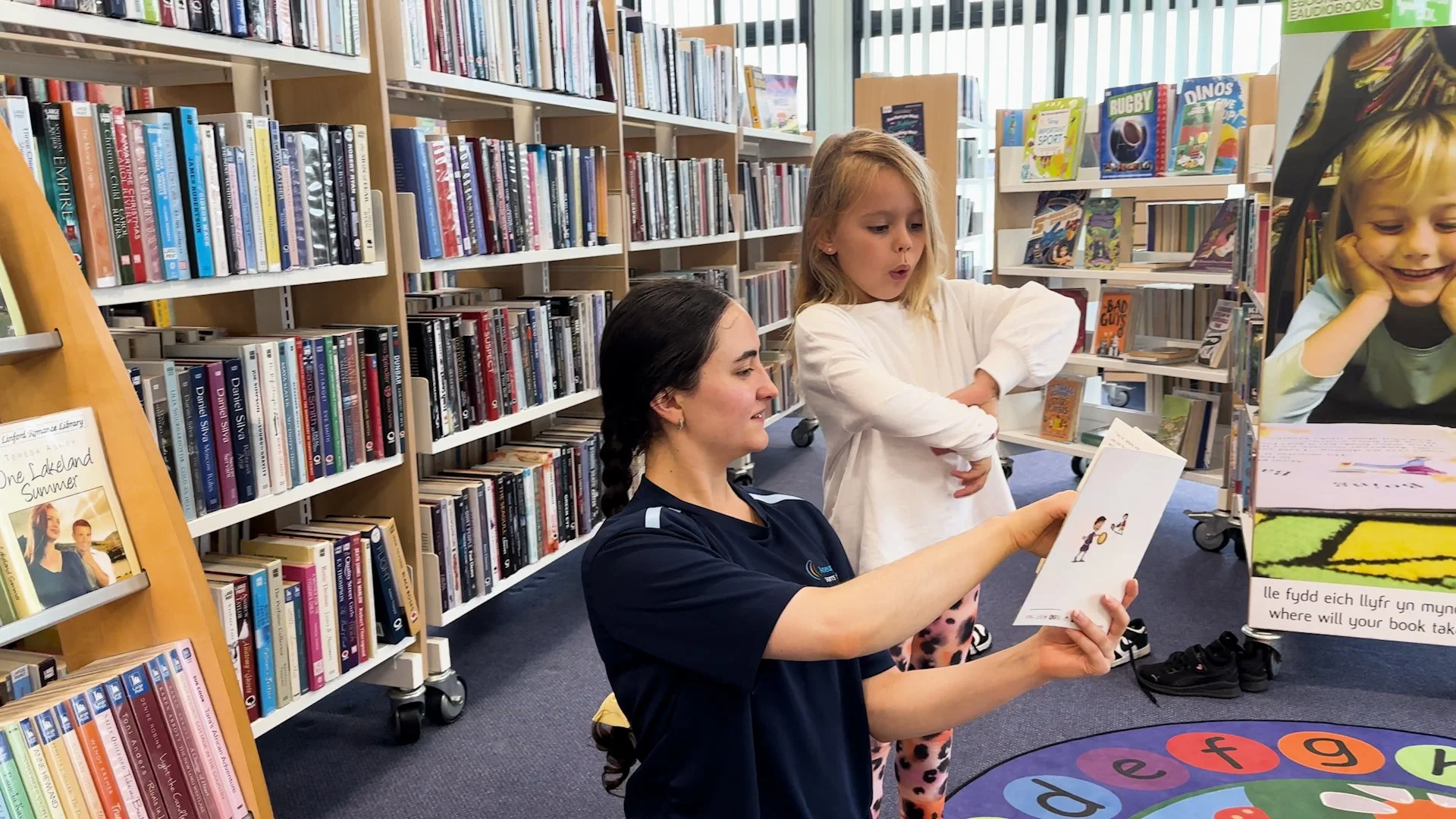 A woman with dark hair in a braid reading a children's book to two young girls in a library, surrounded by bookshelves filled with colorful books.
