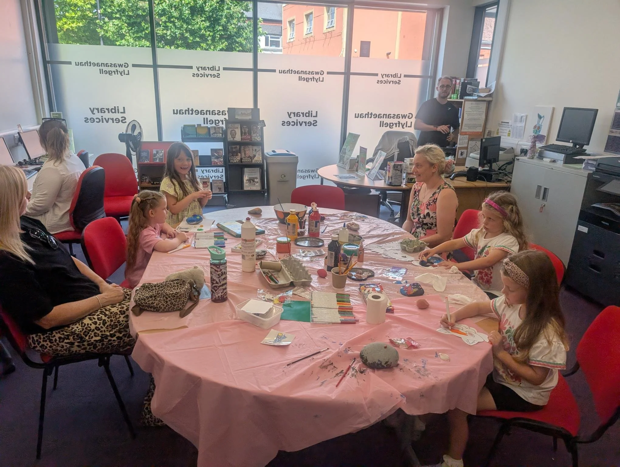Children and adults sitting around a table covered with arts and crafts supplies, engaging in a creative activity at a library event.