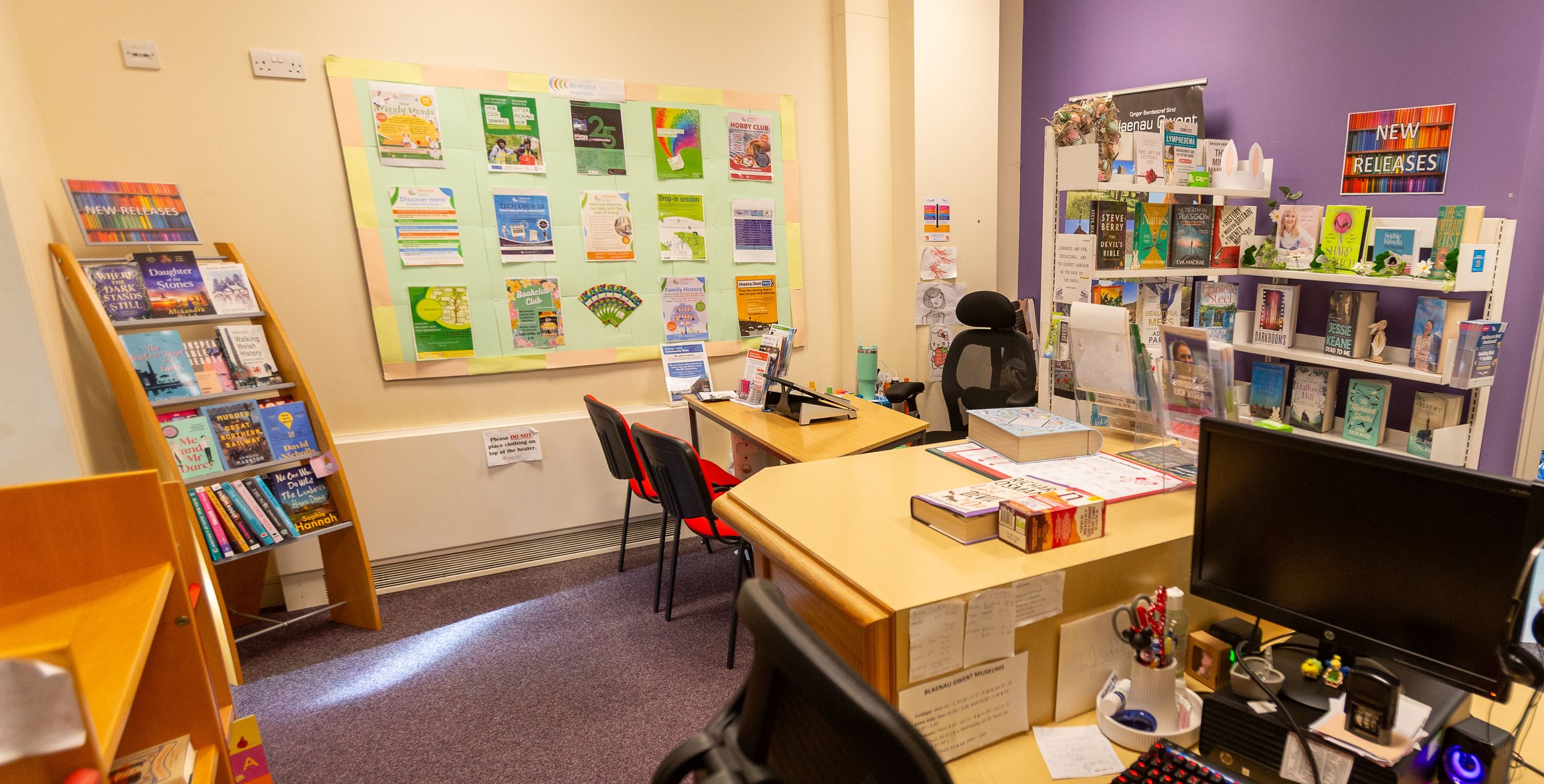 A small library or reading room with bookshelves, a bulletin board, and desks with chairs.