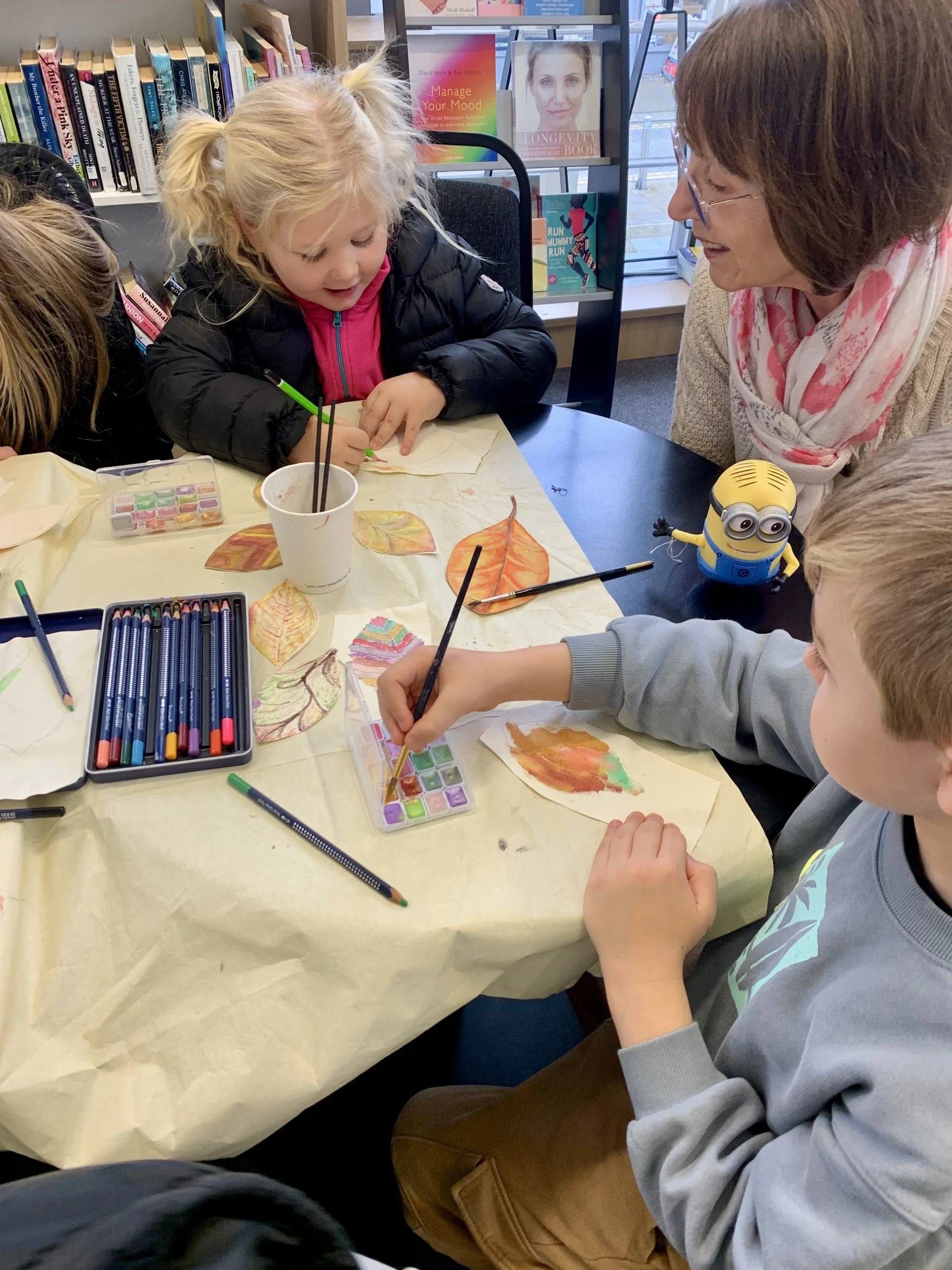 Children and an adult are engaging in an art activity, coloring paper leaves with watercolors and colored pencils at a table in a library.