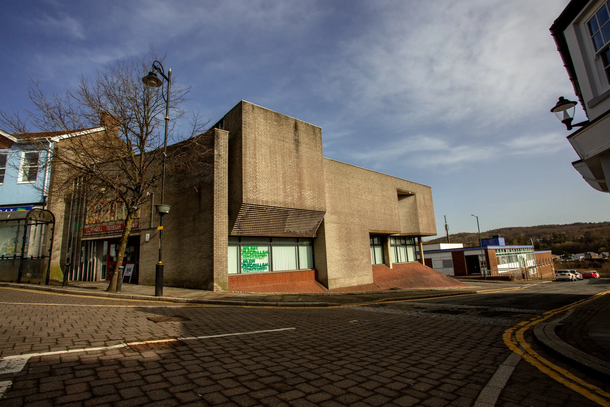 A street corner with a modern brick building, leafless tree, and street lamps under a partly cloudy sky during daytime.