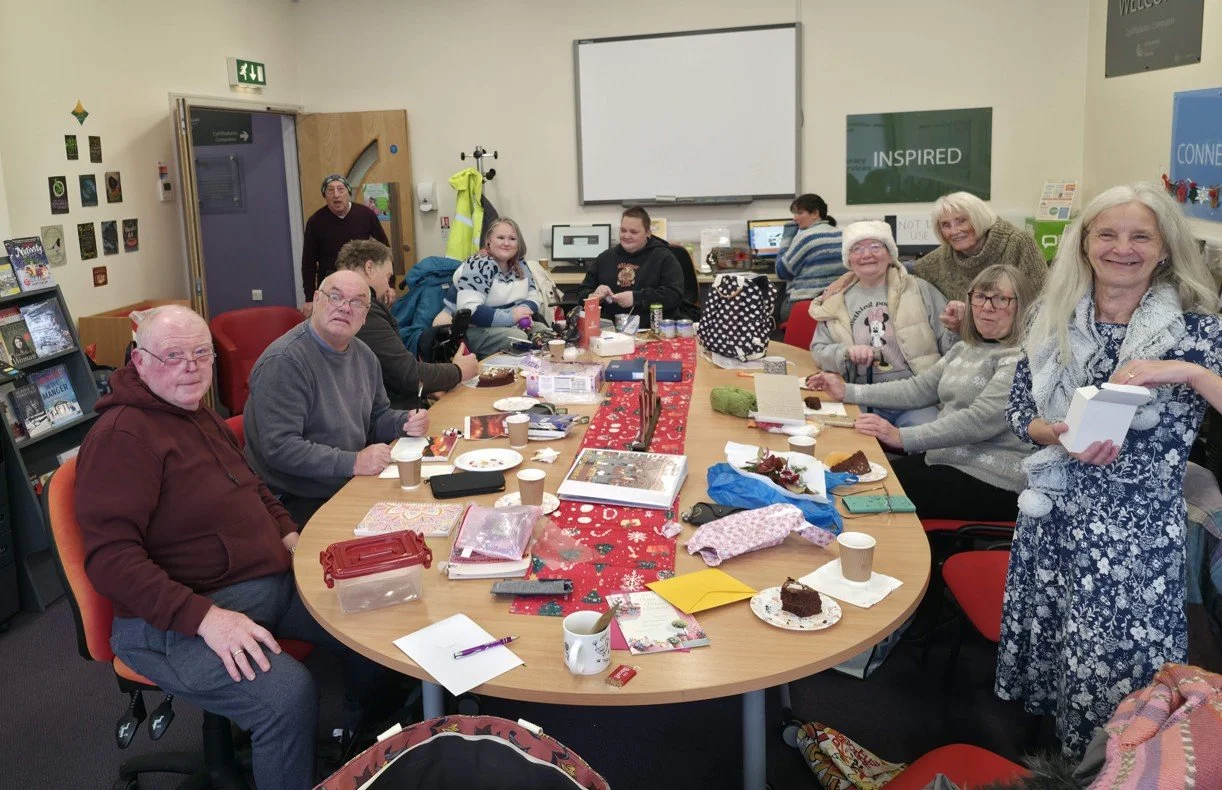 Group of people gathered around a table with Christmas decorations, cakes, and gifts, celebrating in a room with holiday-themed decor.