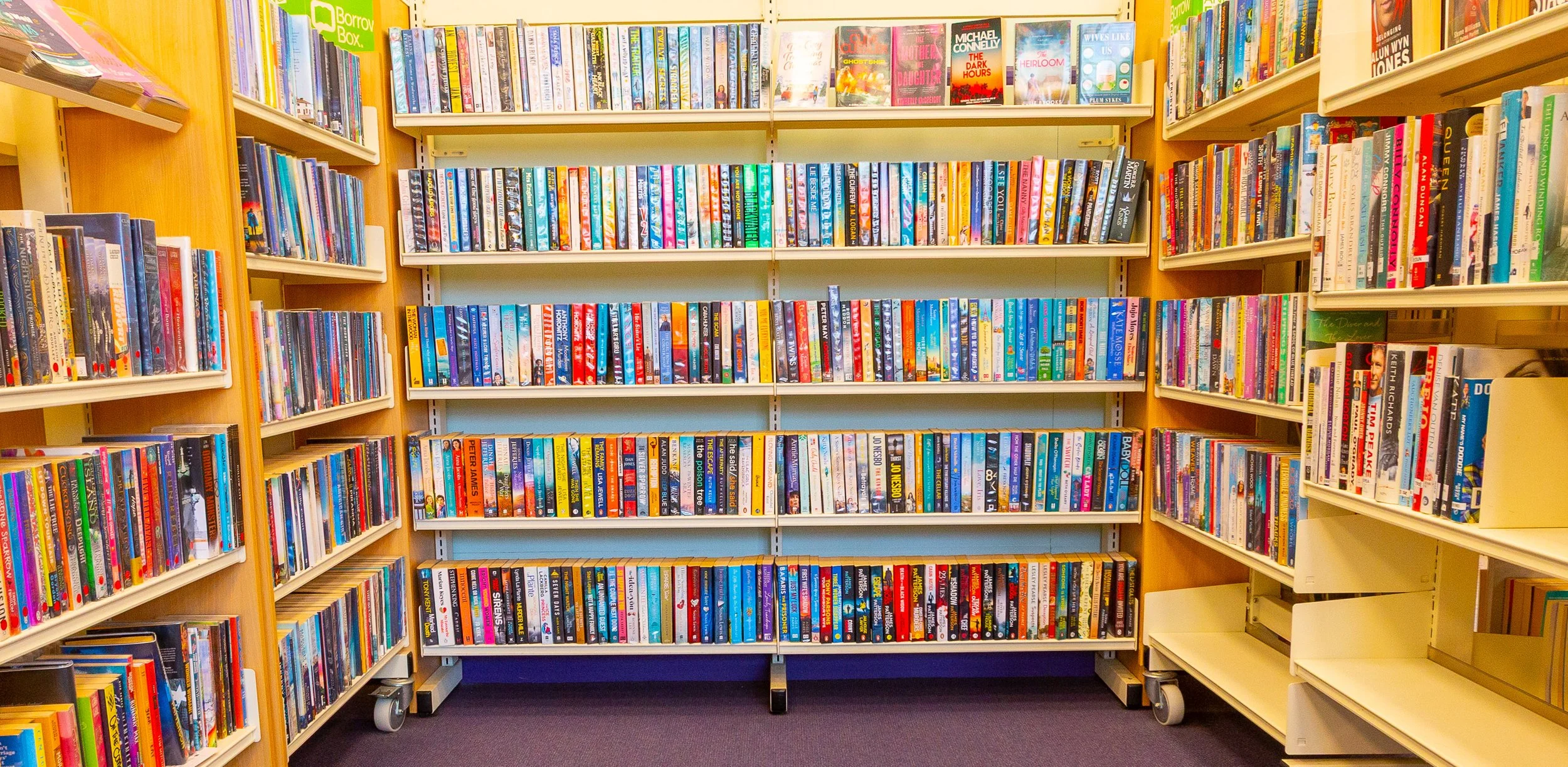 Bookshelves filled with colorful paperback books in a library, with a green Borrow Box sign at the top left.