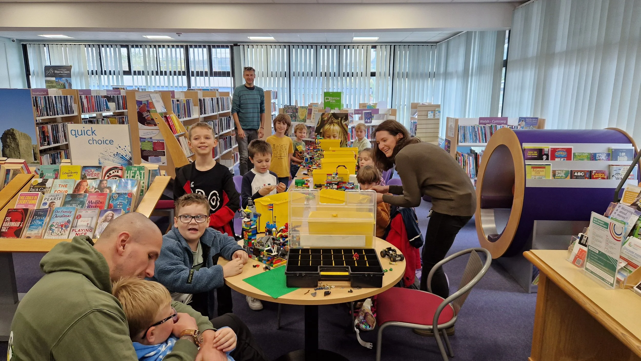 Children and adults in a library participating in a LEGO building activity, with bookshelves, magazines, and seating area visible.
