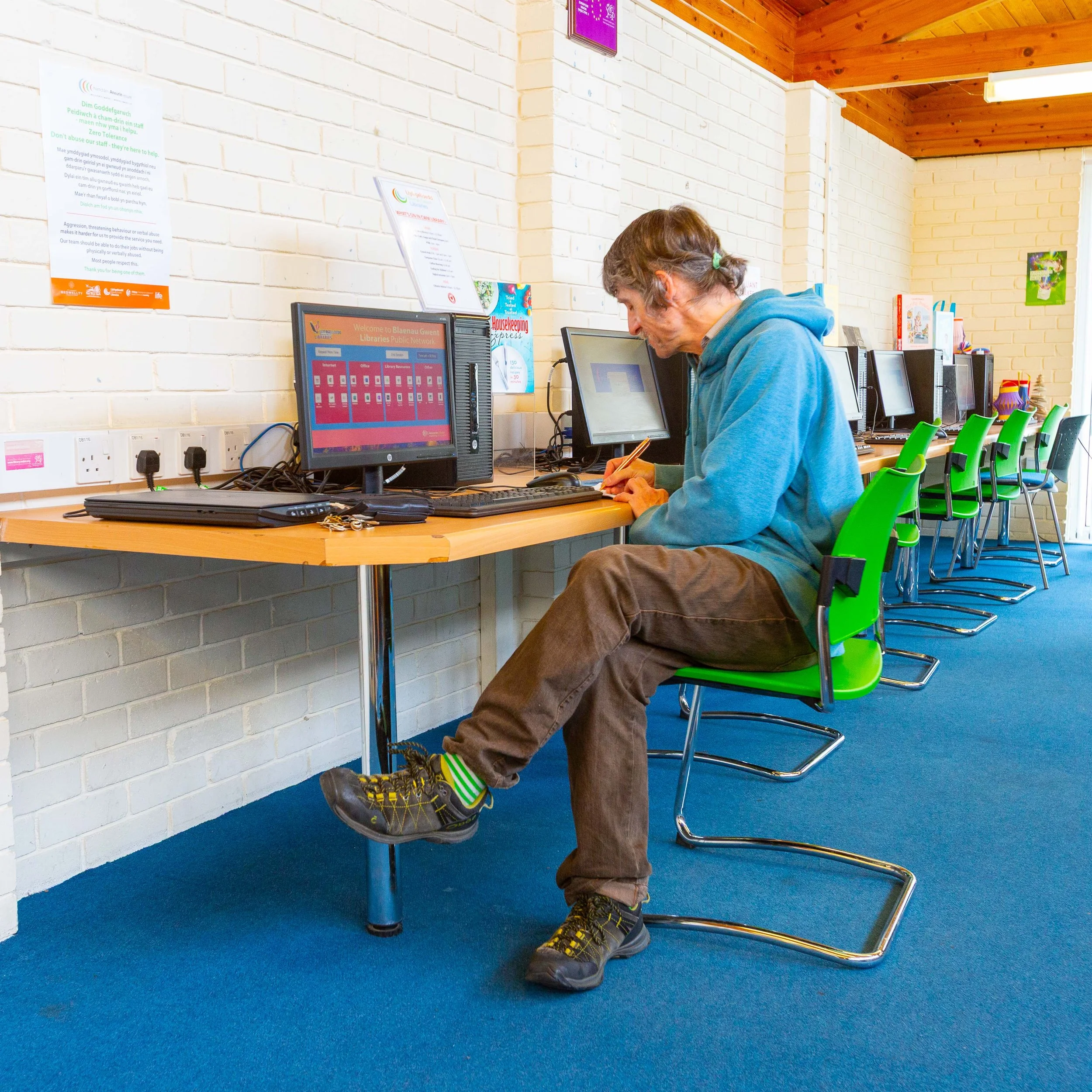 A man in a blue hoodie sitting at a row of green chairs with computers, writing on paper in a library or computer lab with white brick walls and wooden ceiling.