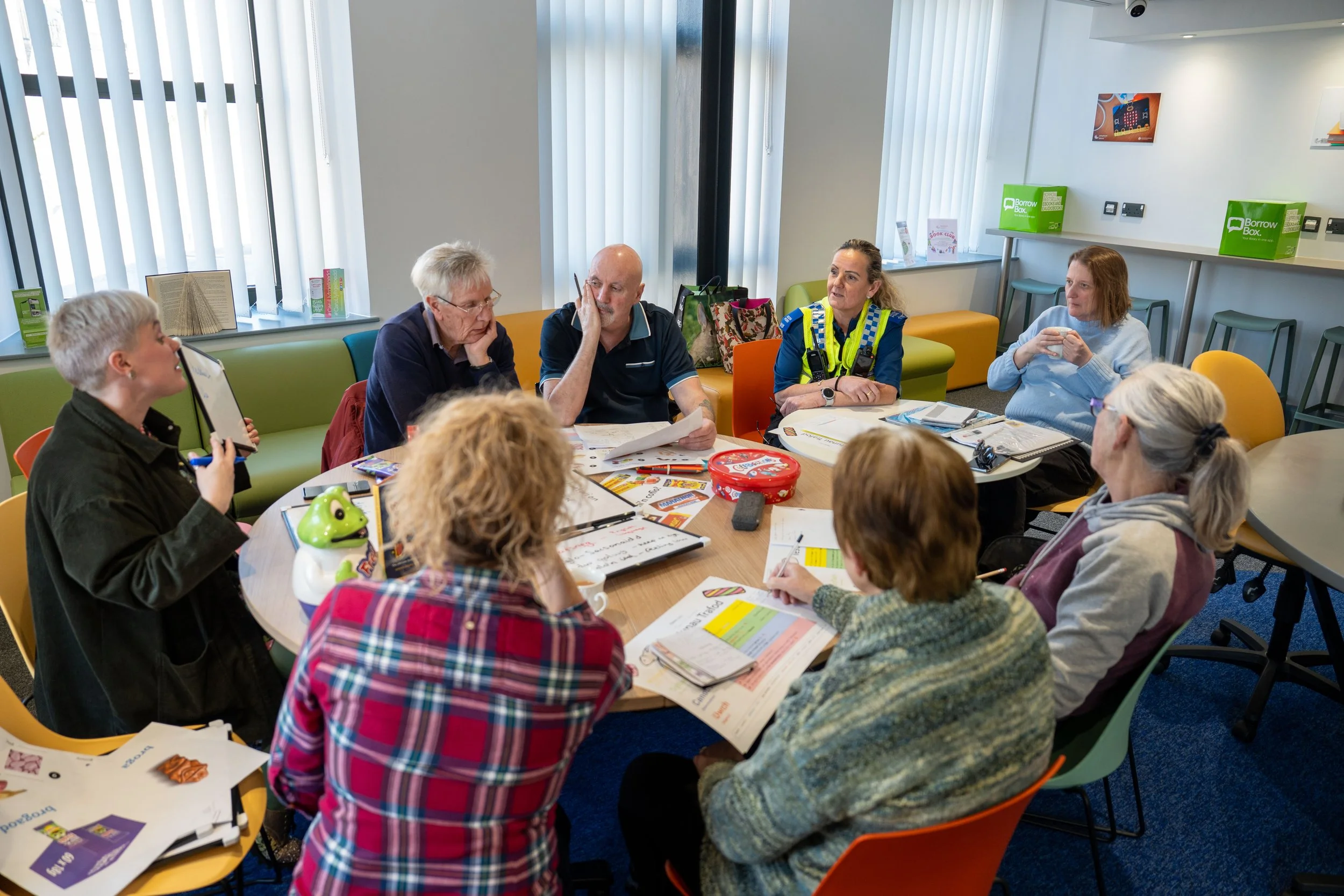Group of nine people sitting around a circular table in a room with large windows, engaging in a discussion or meeting.