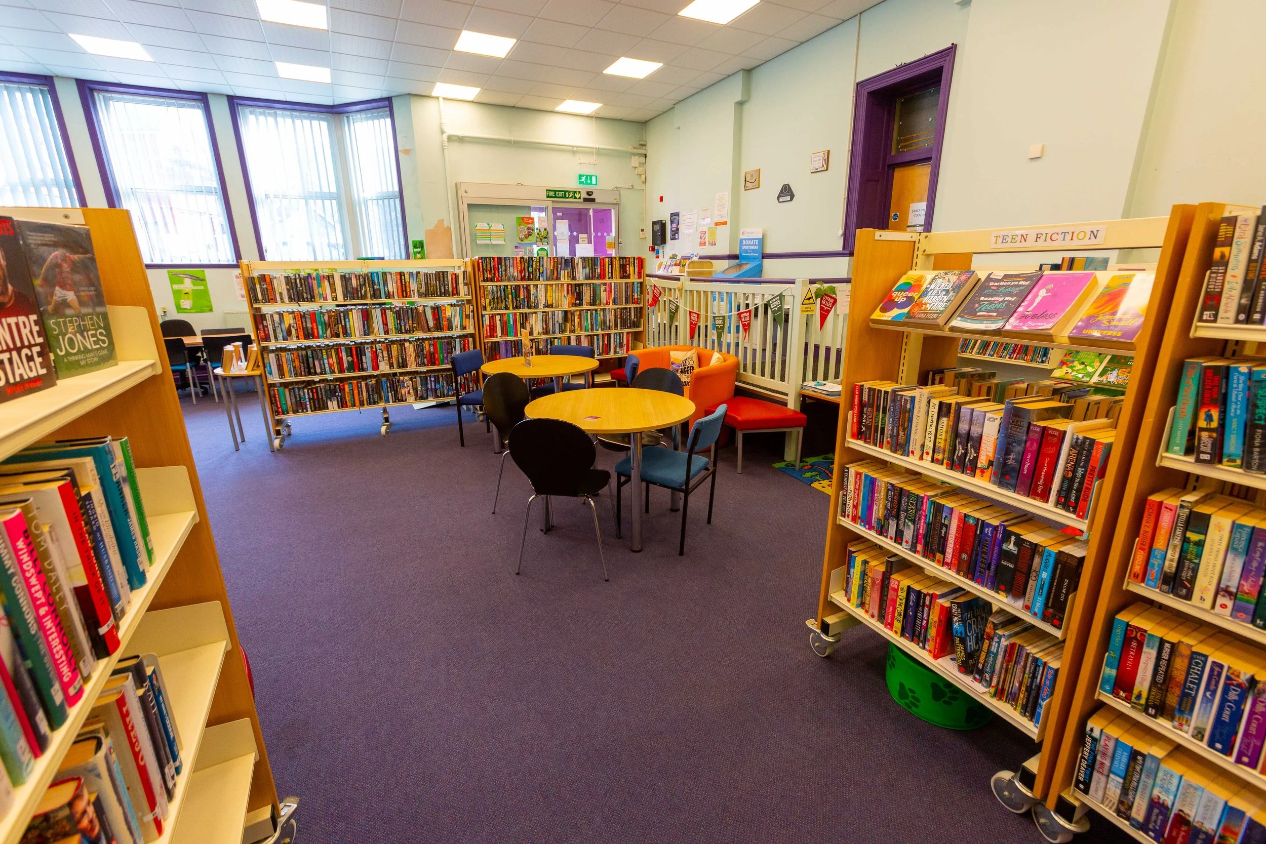 Interior of a library with bookshelves filled with books, small tables and chairs, colorful armchairs, and large windows with blinds.