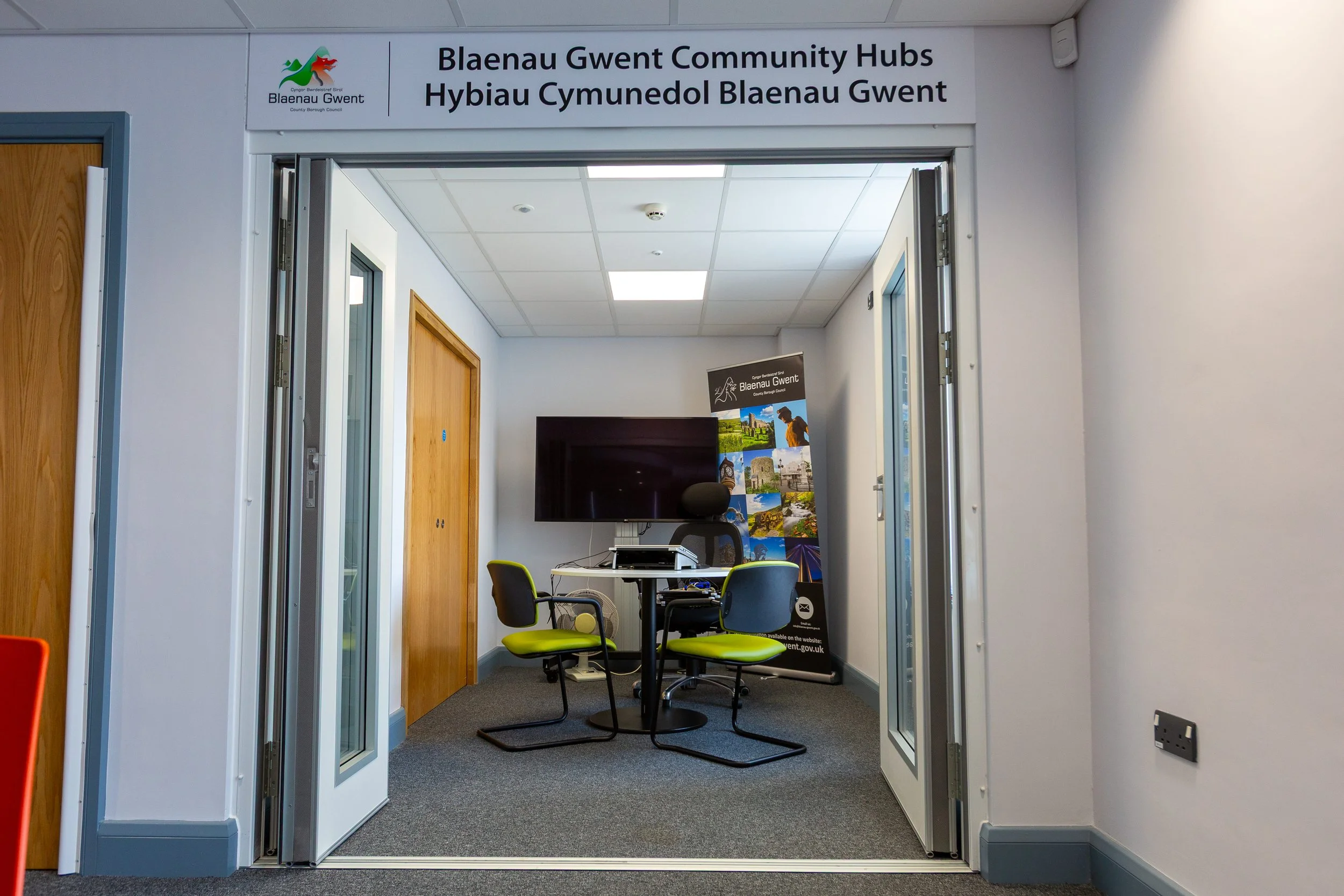 Interior of a community hub office with a large monitor, a table, and three chairs. Signs display Welsh language text and local imagery.