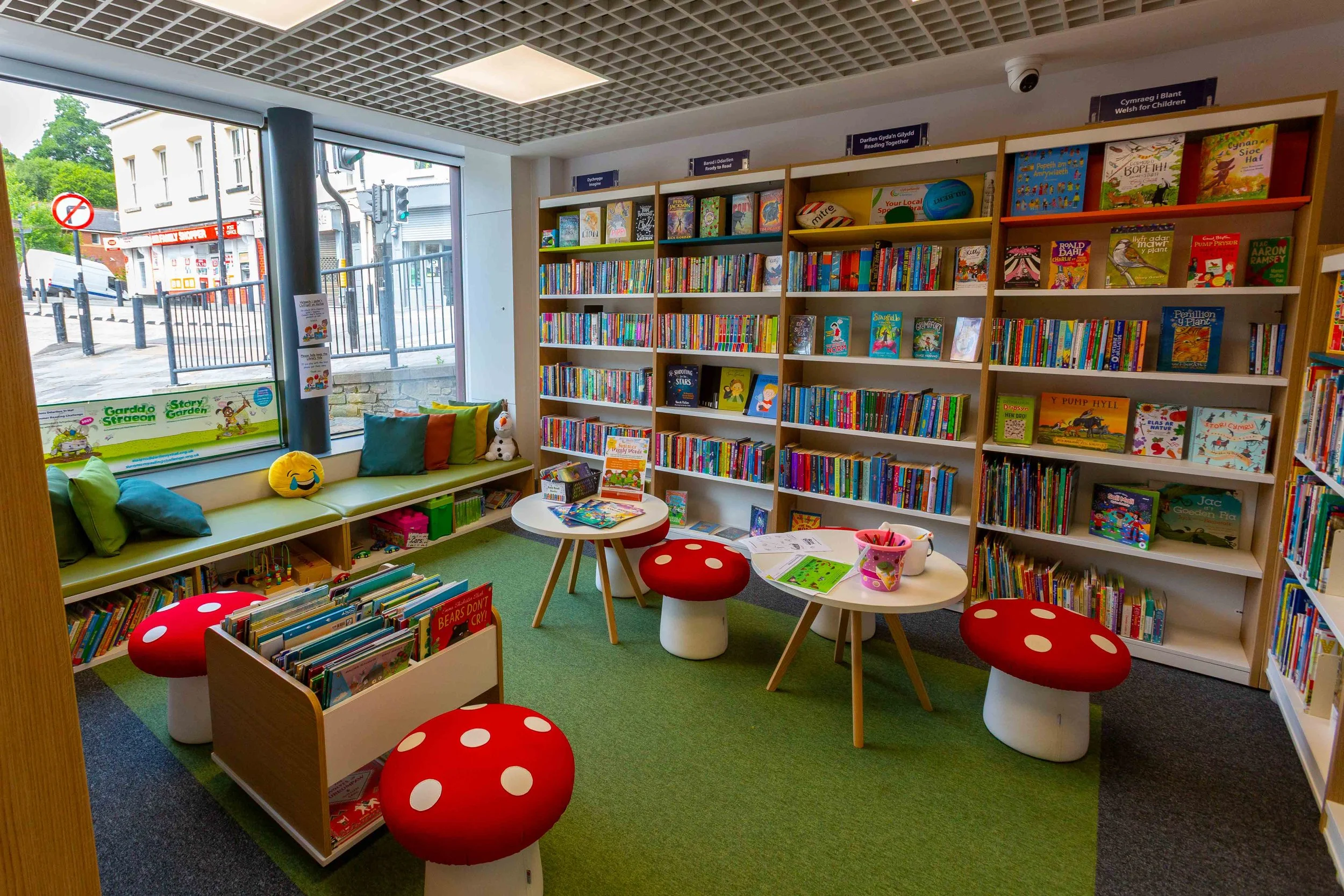 A cozy children's section in a bookstore or library with colorful bookshelves filled with children's books, a window seat with plush toys and cushions, round tables with red mushroom-shaped stools, and a rug in shades of green and gray.