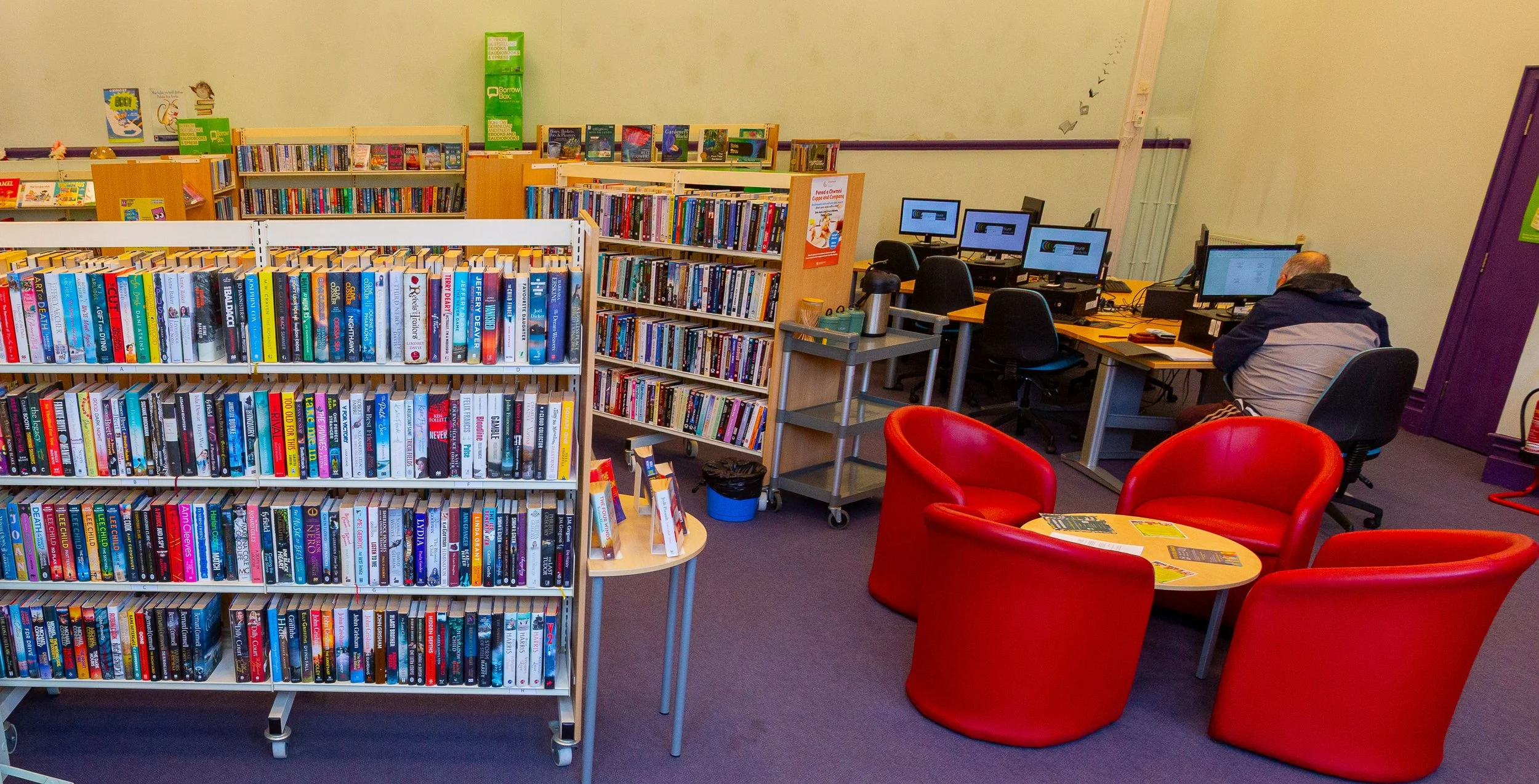 A library or computer room with bookshelves filled with books, a group of four red chairs arranged around a small table with magazines, and four computer workstations with people working at them.