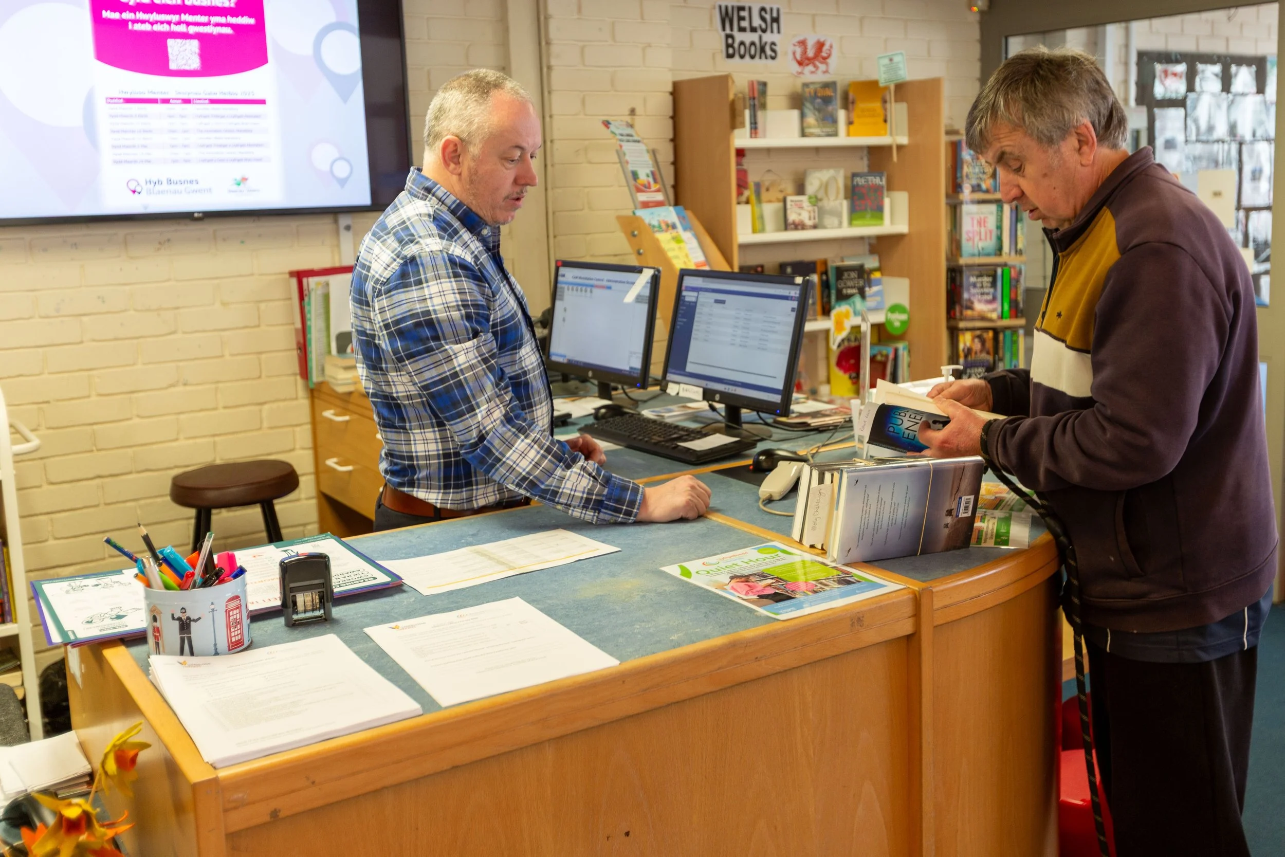 A man at the counter of a bookstore or library with a cashier wearing a plaid shirt, assisting a customer who is reading a book. Shelves filled with books and a large screen displaying information are visible in the background.
