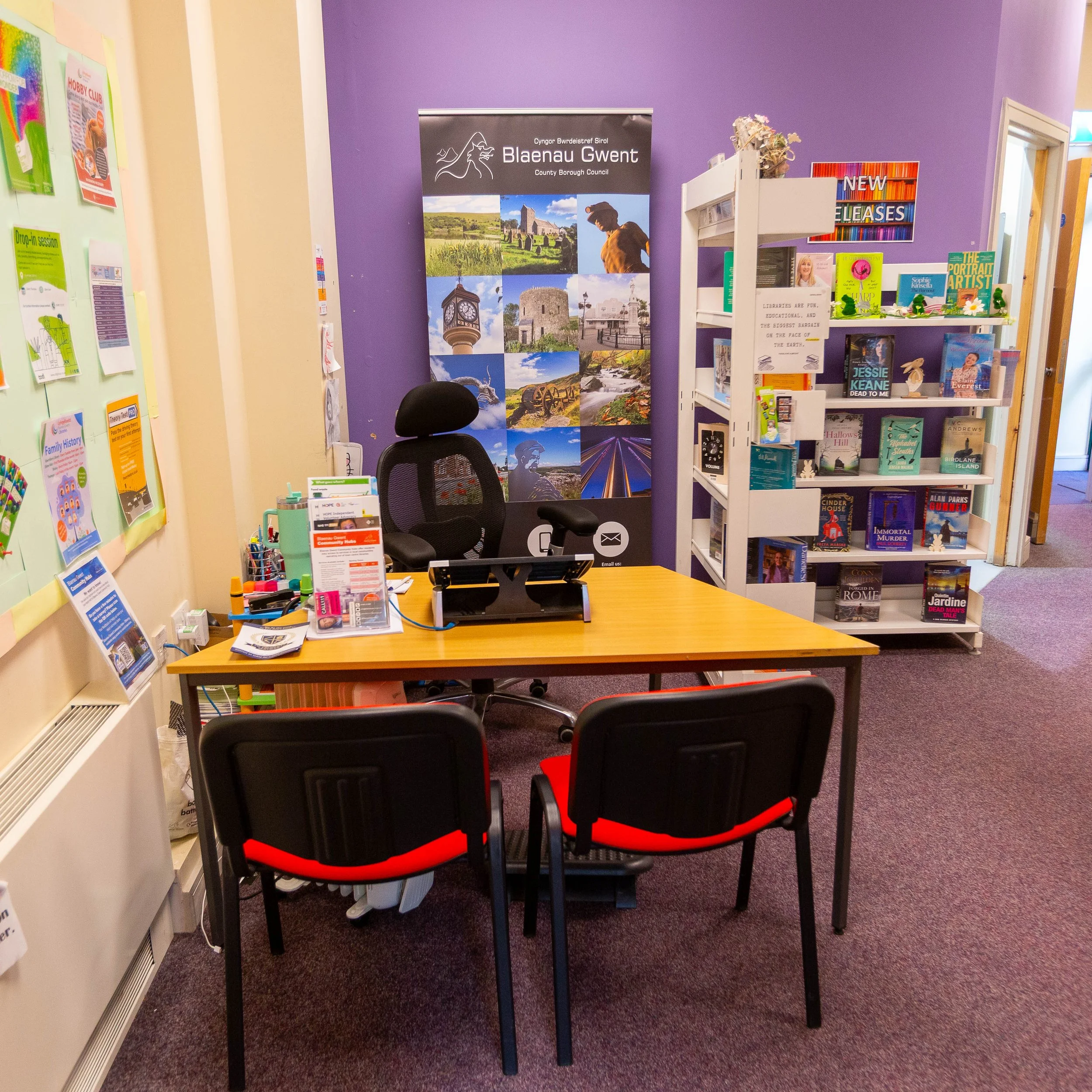 An office reception desk area with black chairs, a wooden table, a black swivel chair behind the desk, and a purple wall with a Blaenau Gwent Council poster showing images of local landmarks. A white bookshelf on the right displays books and magazine