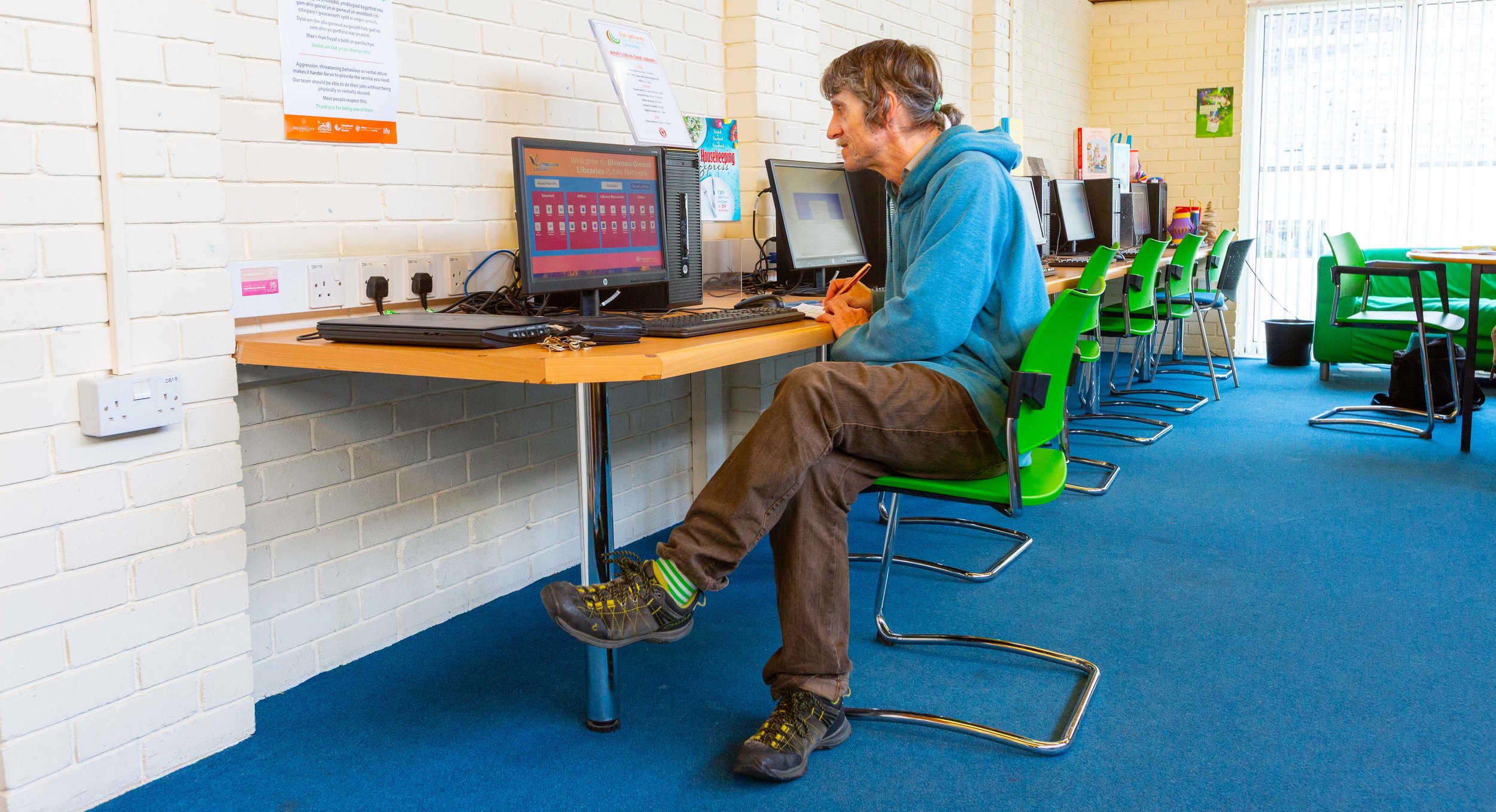 A man in a blue hoodie and brown pants working on a computer in a room with green chairs and a blue carpet.