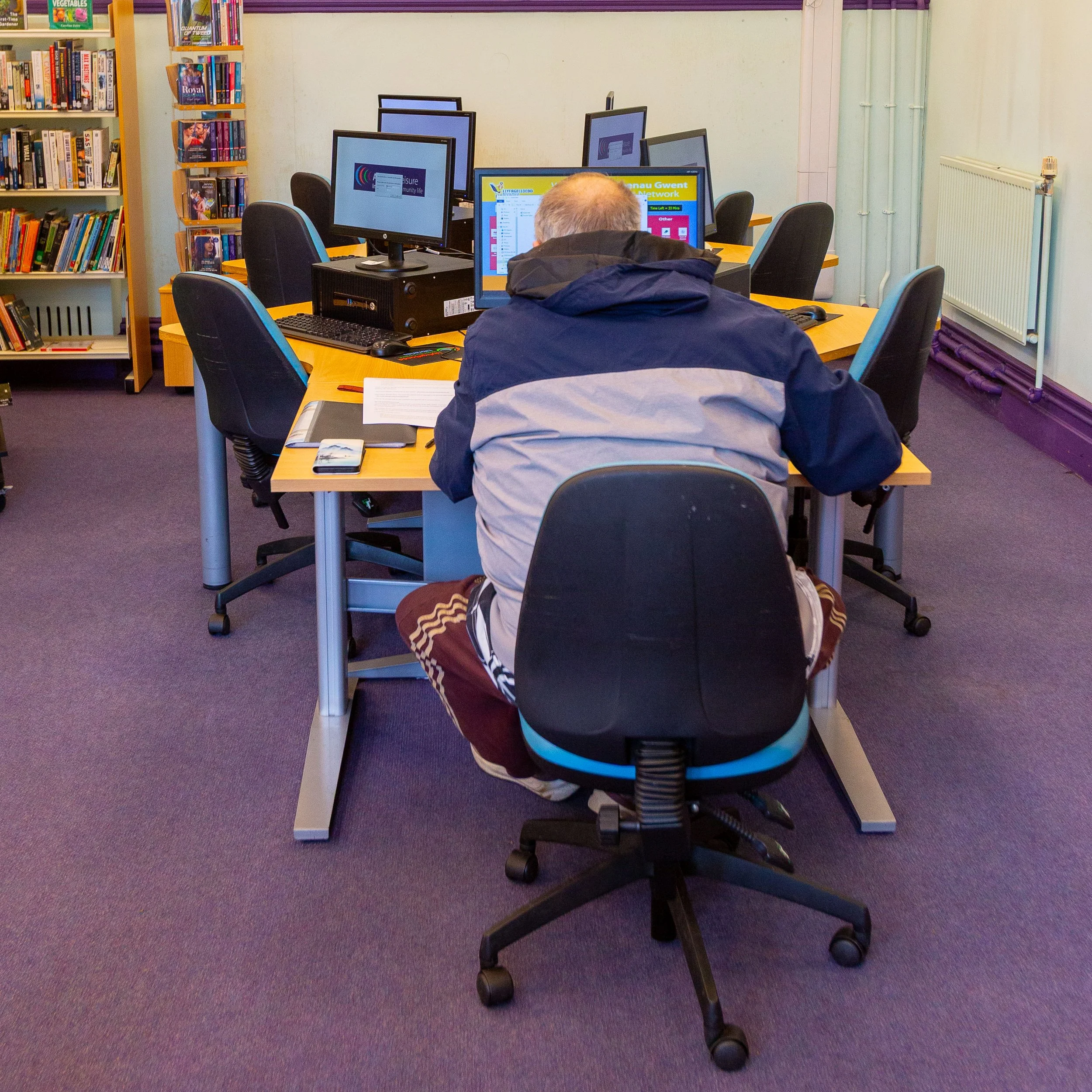 A person wearing a jacket and sweatpants sitting at a computer desk in a library or computer lab, with multiple monitors and bookshelves in the background.