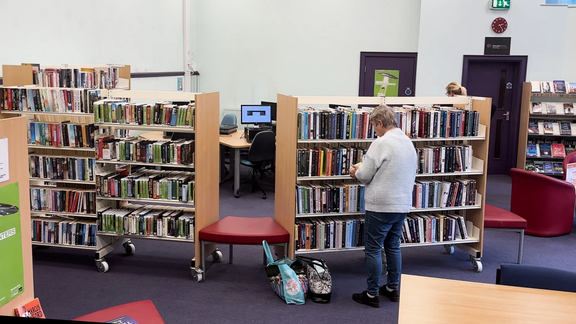 A man browsing books in a library or bookstore, with bookshelves, a computer, and chairs in the background.