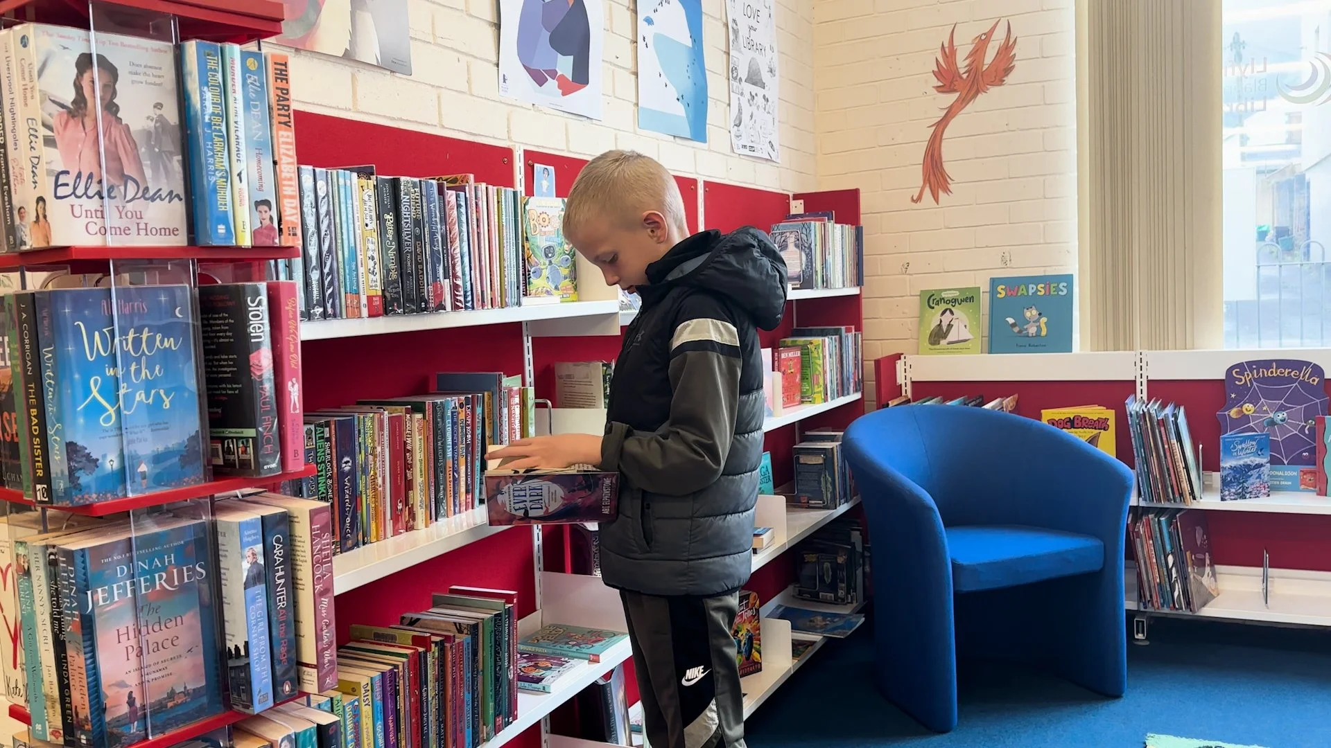 A young boy reads a book in a cozy library corner with red and white bookshelves filled with colorful books, a blue armchair, and children's artwork on the yellow brick wall.