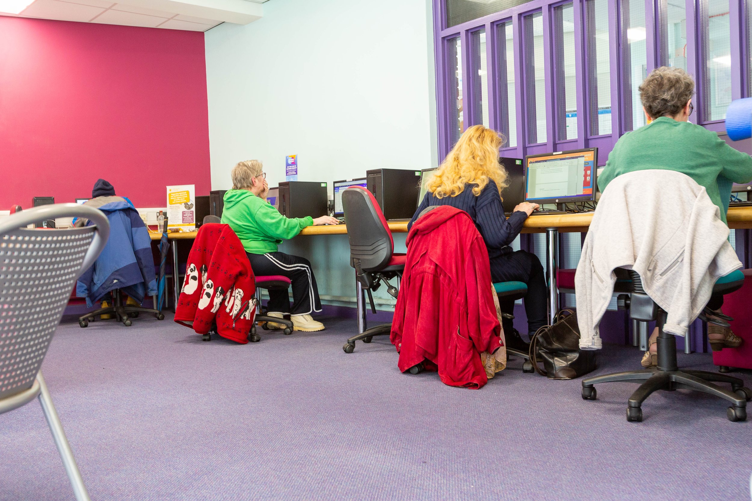 People using computers in a colorful public library or community center with purple window frames and a pink accent wall.