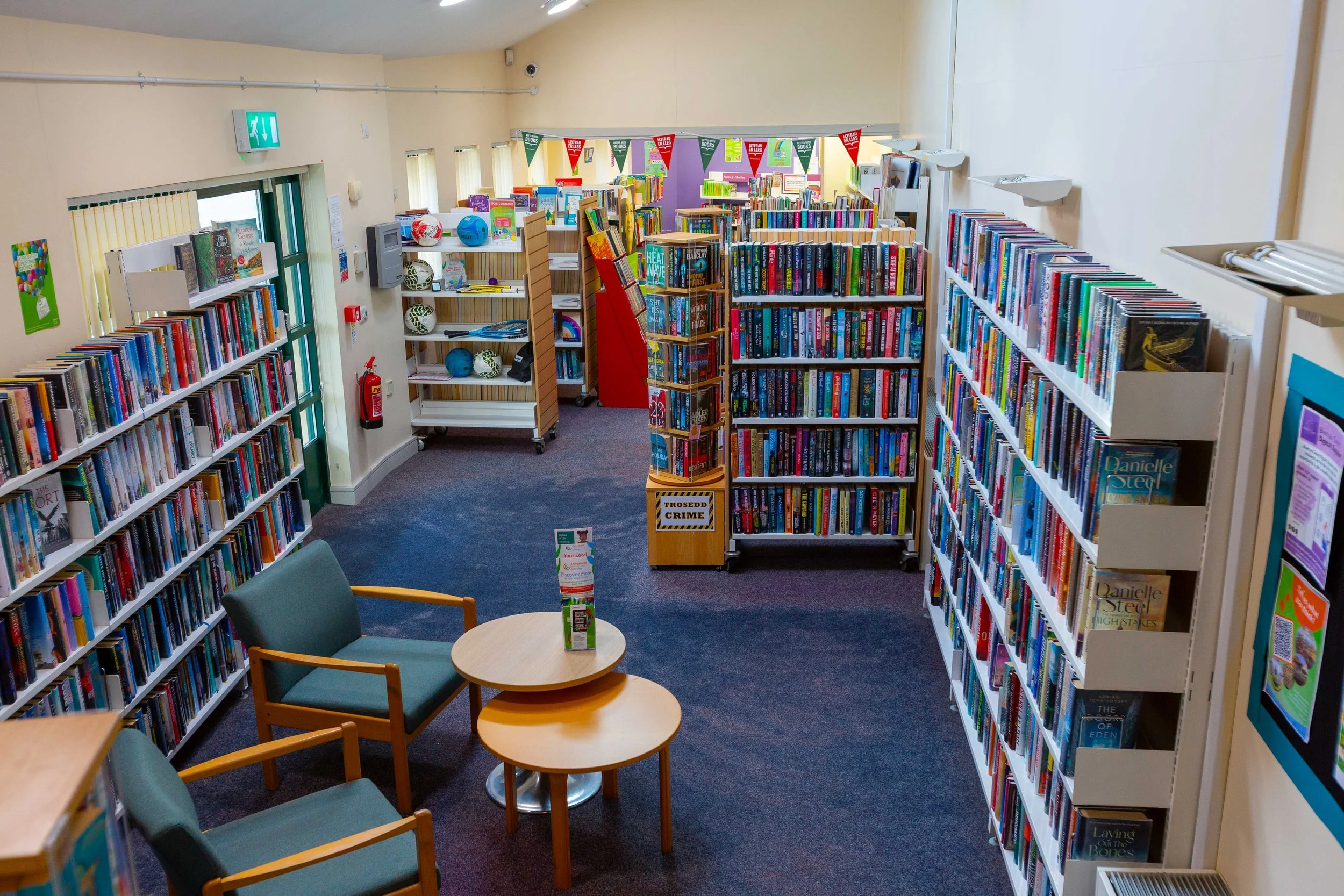 Interior of a small library with shelves filled with books, some chairs and tables for reading, and colorful bunting hanging from the ceiling.