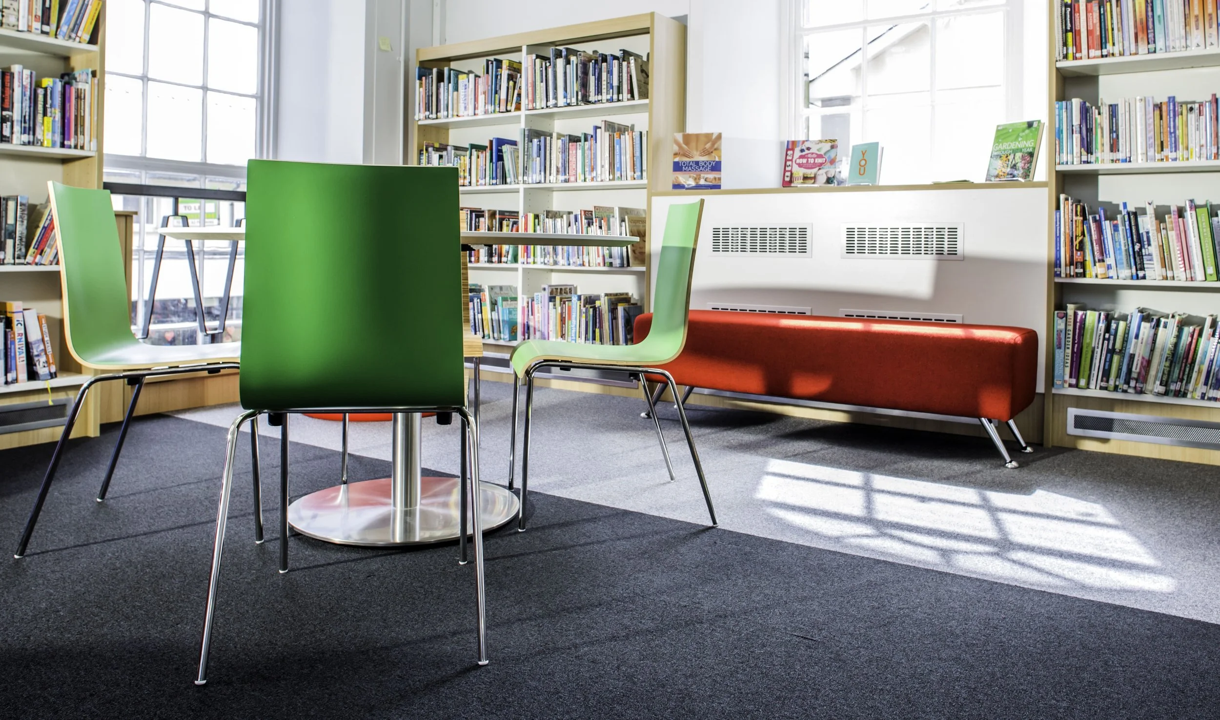 Interior of a library with bookshelves, green and beige chairs, and a red bench. Sunlight streams through large windows.