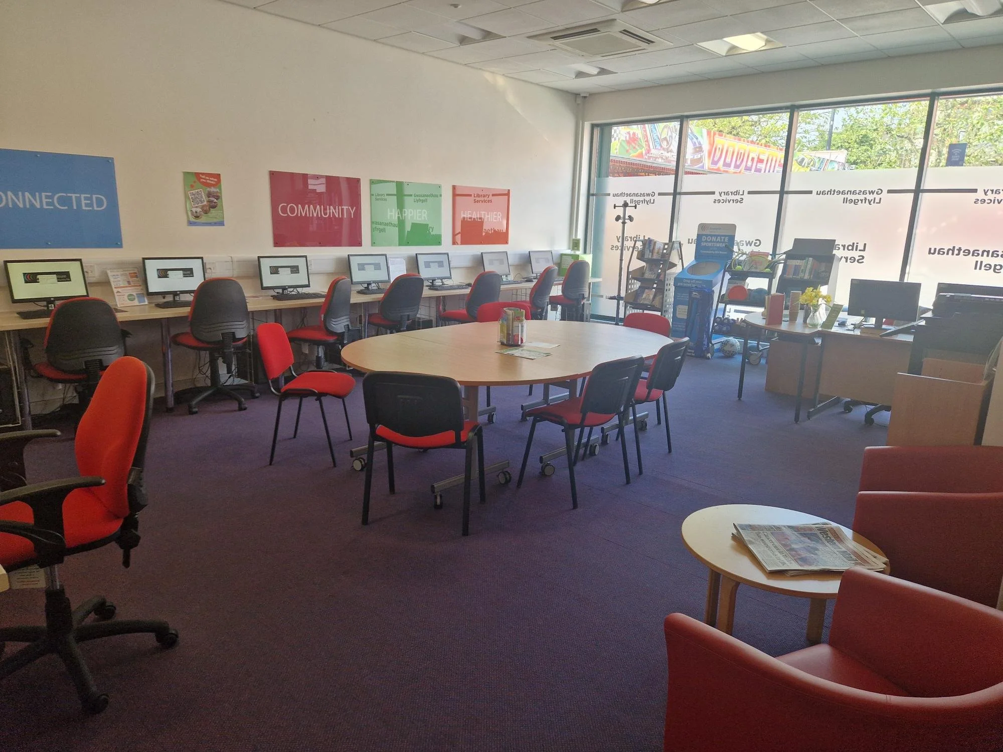 A computer room with multiple desks and chairs, some facing computer monitors, and posters on the wall reading 'Connected,' 'Community,' 'Happier,' and 'Healthier.' There is a large window with a view outside, a round table in the center, and some ne