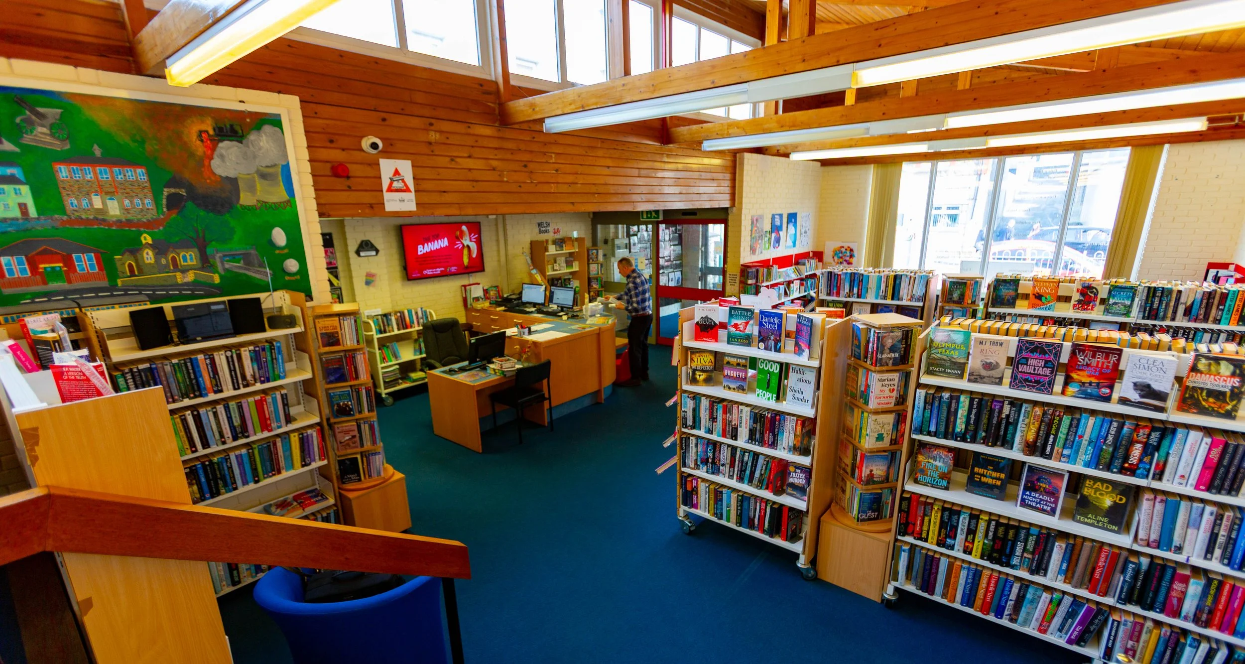 Interior of a bookstore or library with multiple bookshelves filled with books, a wooden ceiling with large windows, a man working at a desk, colorful artwork on the walls, a digital screen displaying 'BANANA,' and a doorway leading outside.