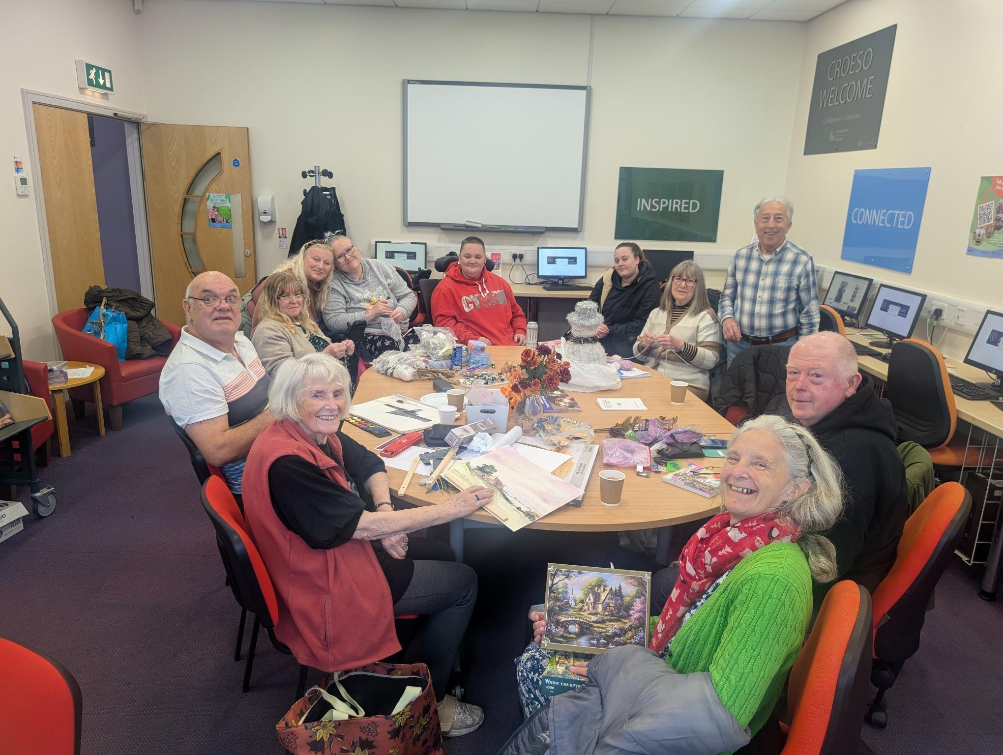 A group of people gathered around a table in a room decorated for Christmas, with some holding gifts and a small decorated Christmas tree in the center. They are smiling and appear to be celebrating.