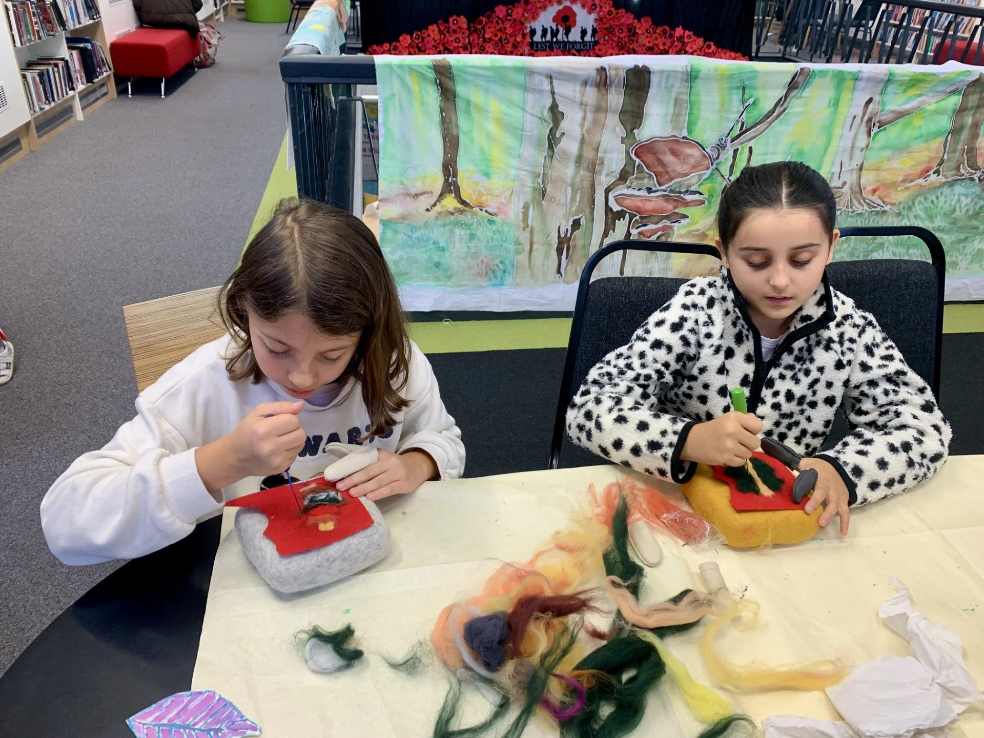 Two young girls sitting at a table, working on felting craft projects with colorful wool. There are felting tools in their hands, and materials are spread out on the table. A painted backdrop featuring a forest scene is behind them.
