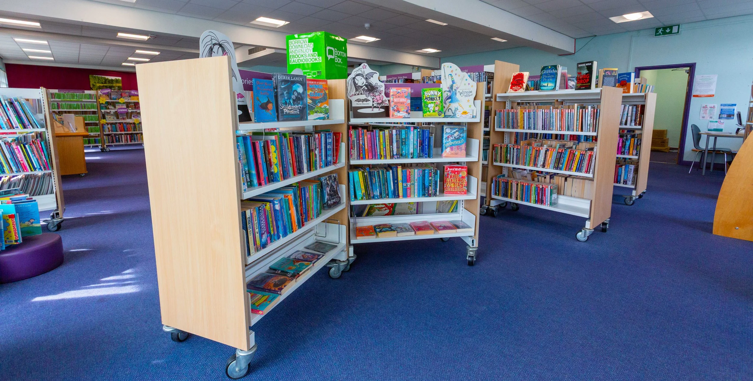 Inside a library with several bookshelves filled with children's books on blue carpeted floor.