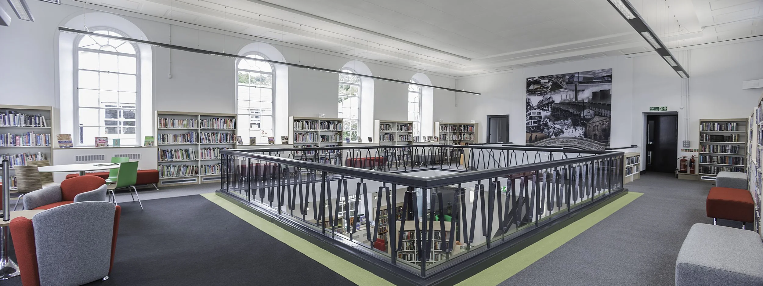 A modern library interior with white walls, large arched windows, filled bookshelves, and a black metal railing overlooking a lower level.