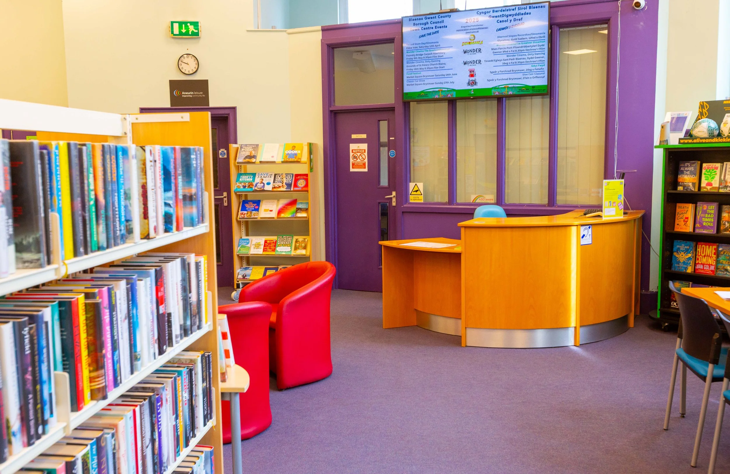 Interior of a colorful library or community center, with bookshelves filled with books, a red chair, and a reception desk with a blue chair. There are purple walls and a large digital display screen with event information.