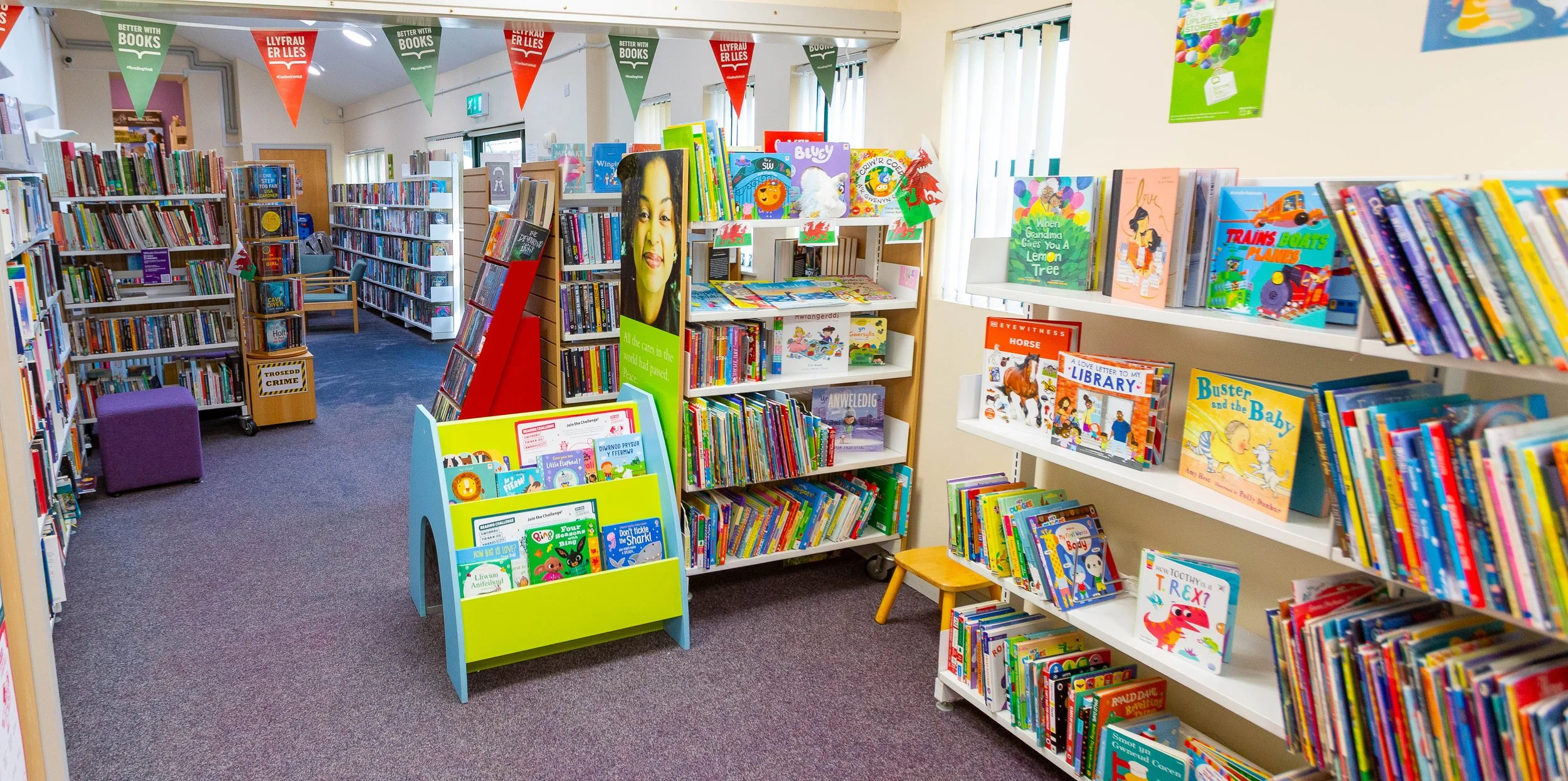 The image shows the interior of a colorful children's section in a library, with bookshelves filled with children's books, a display stand with picture books, and banners overhead promoting reading. The room is well-lit with natural light coming from