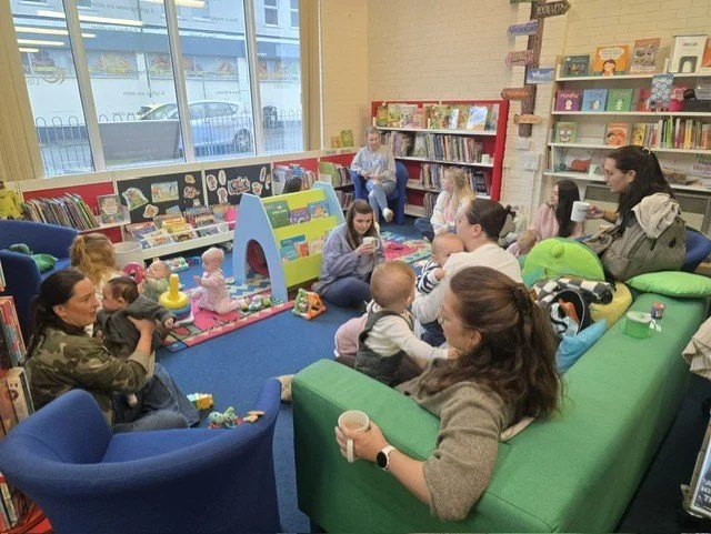 Children and adults sitting in a cozy children's library or bookstore. Shelves filled with colorful books line the walls, and a woman in a camouflage jacket is playing with a baby among stuffed toys on the floor. Others are sitting on a green couch, 