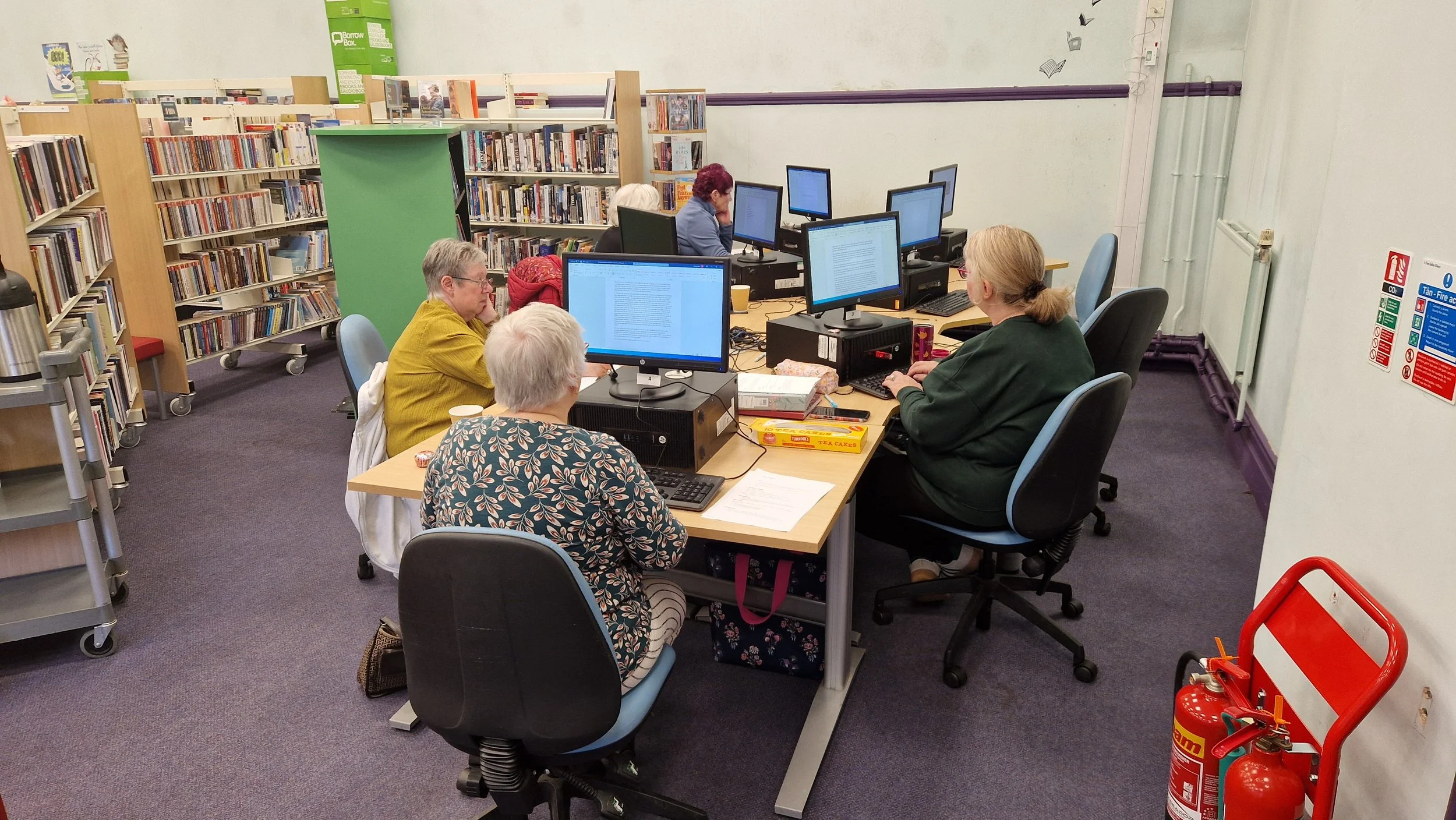 A group of five elderly women working on computers in a library, surrounded by bookshelves, with a fire extinguisher and safety signs in the corner.