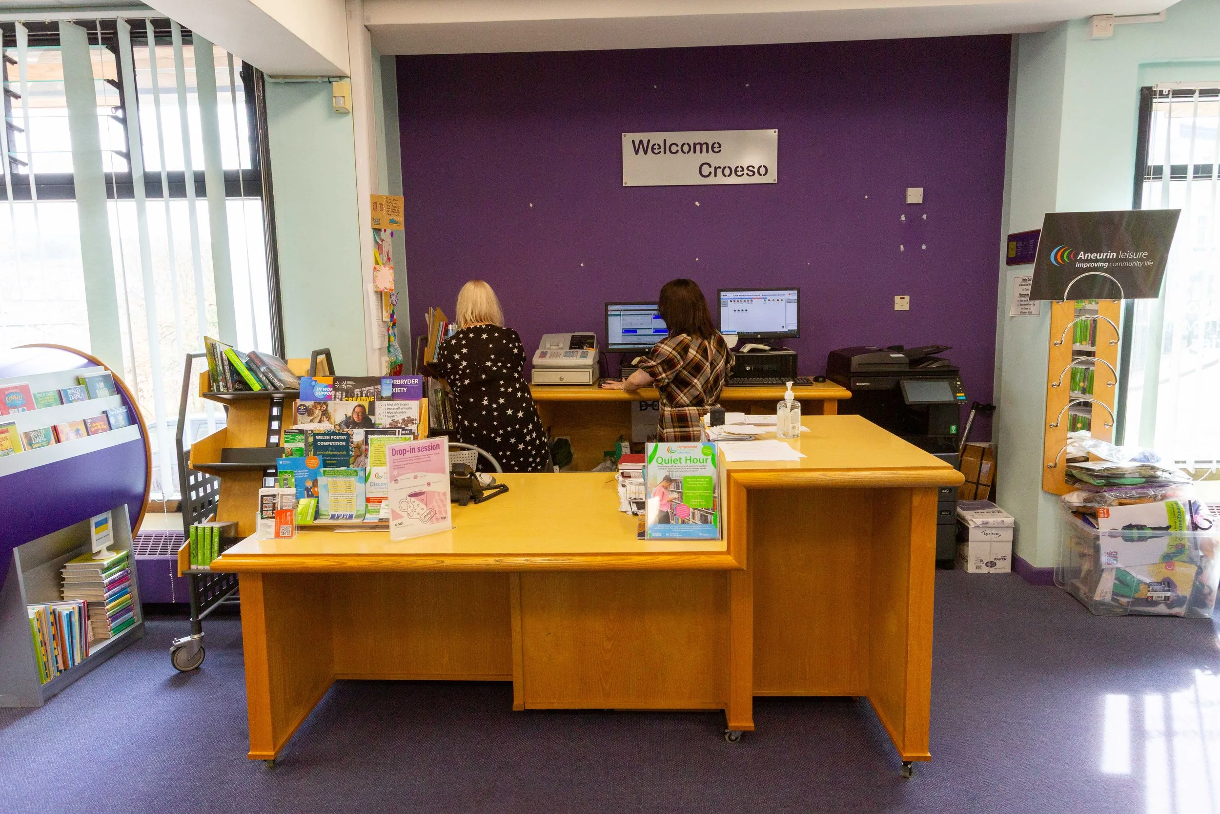 Reception area of a library or community center with two women working at computers. There are books and brochure stands on the desk and shelves, a sign saying 'Welcome Croeso' on the purple wall, and windows allowing sunlight into the room.