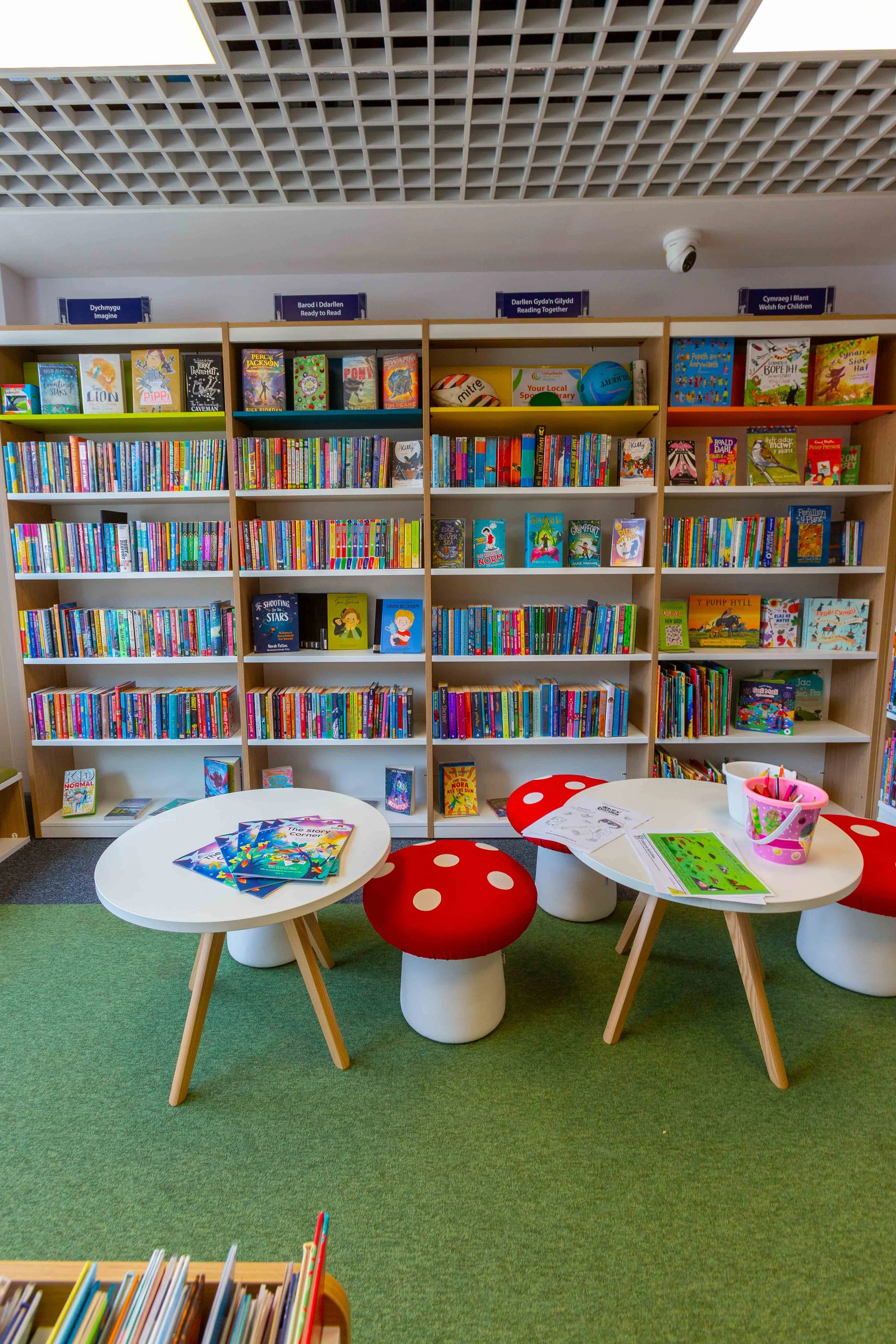 Children's section of a library with bookshelves full of colorful books, two white round tables with children's books and coloring pages, red mushroom-shaped stools, and green carpet.
