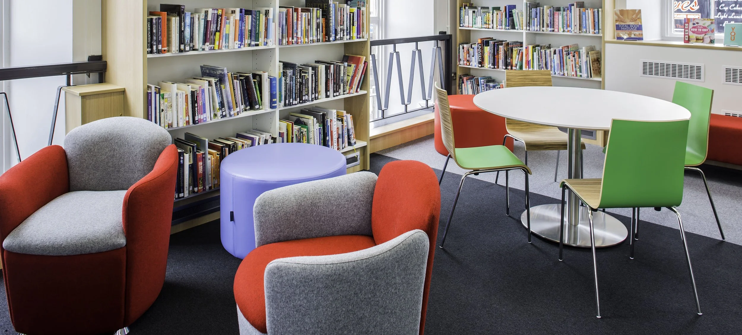 A cozy library or reading area with bookshelves filled with books, colorful chairs, and round tables, illuminated by natural light from windows.