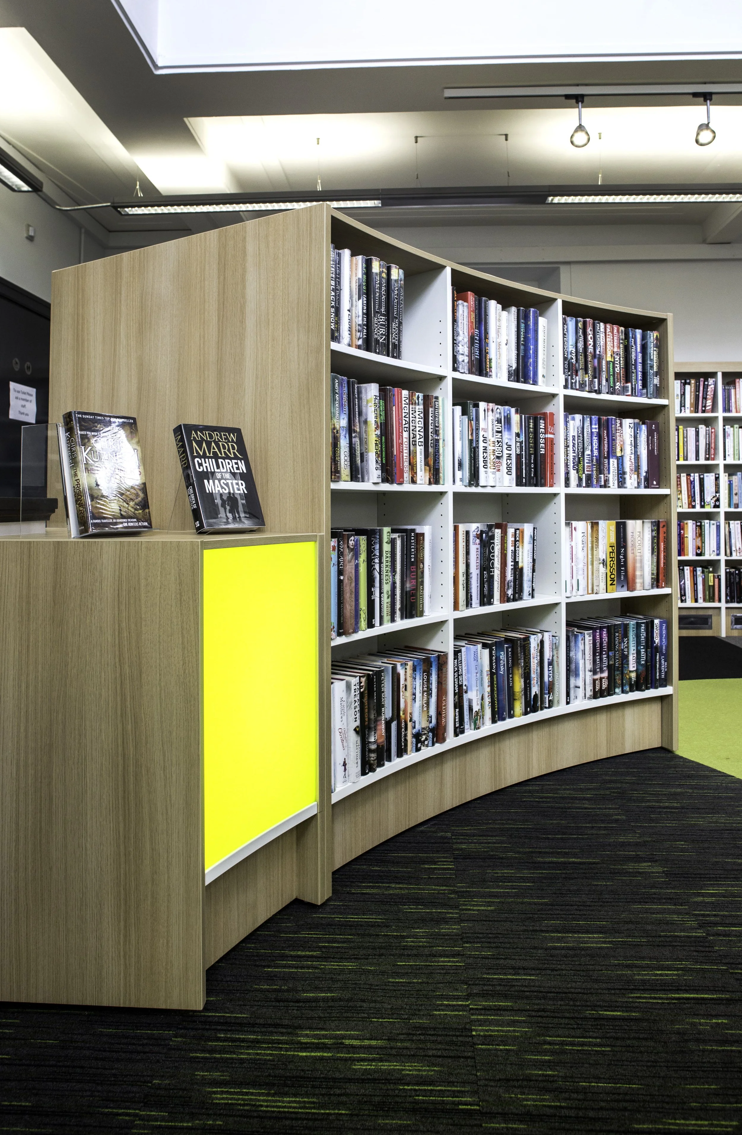Curved bookshelf filled with books in a library, with a yellow panel on the side and a black carpet with green lines.
