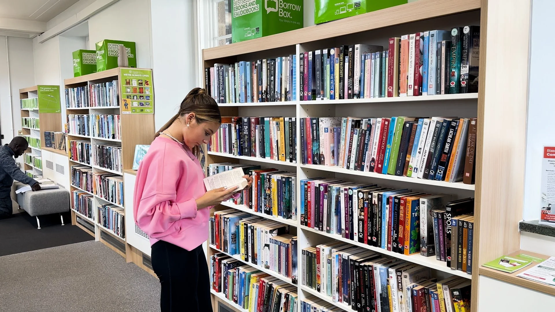 A woman in a pink sweatshirt reading a book in front of a bookshelf in a library. A man is in the background, browsing books.