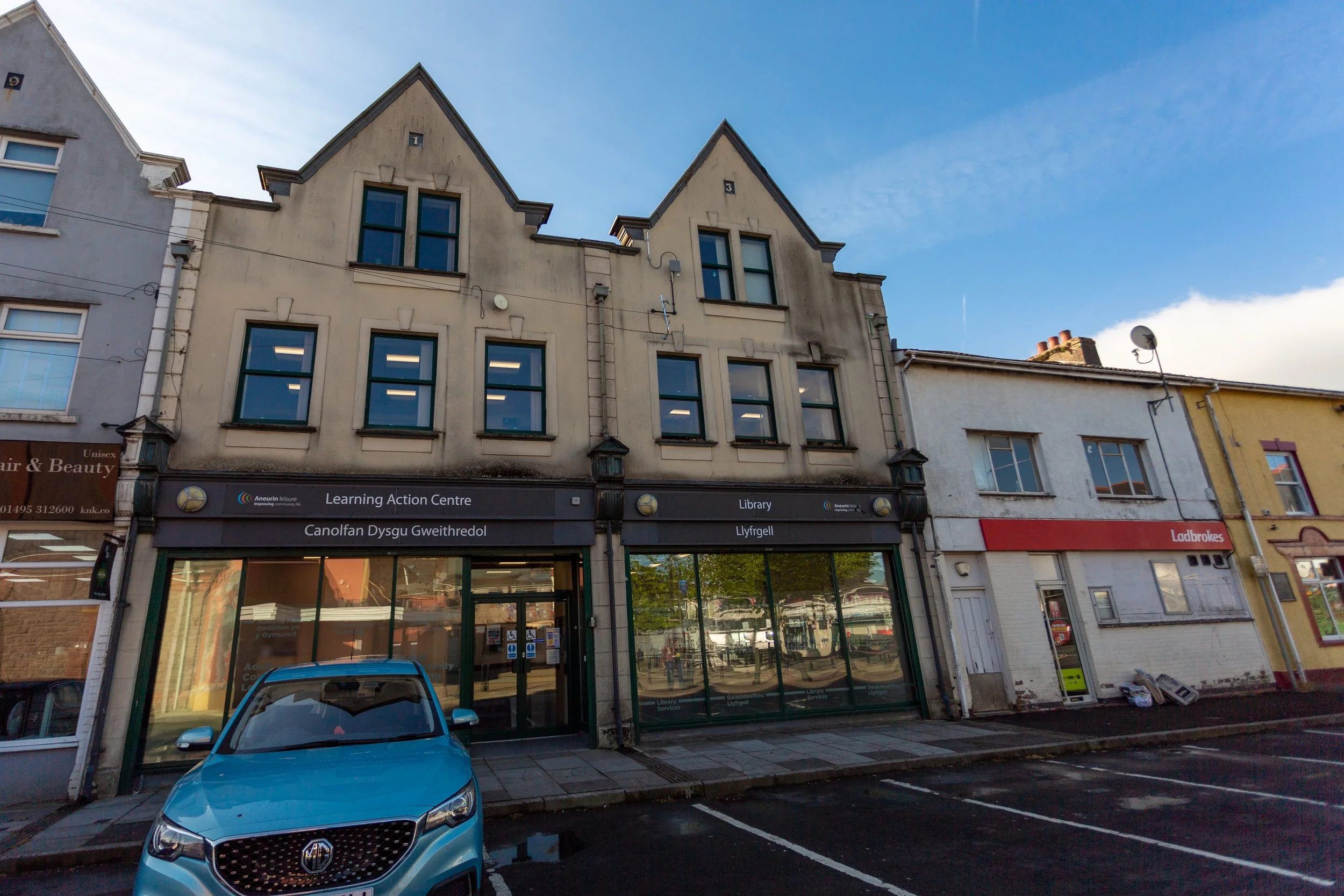 A street view featuring a three-story building with a bookstore on the ground floor, parking lot in front, and a blue car parked. The building has gabled windows on each level, and signage for the library and learning center.