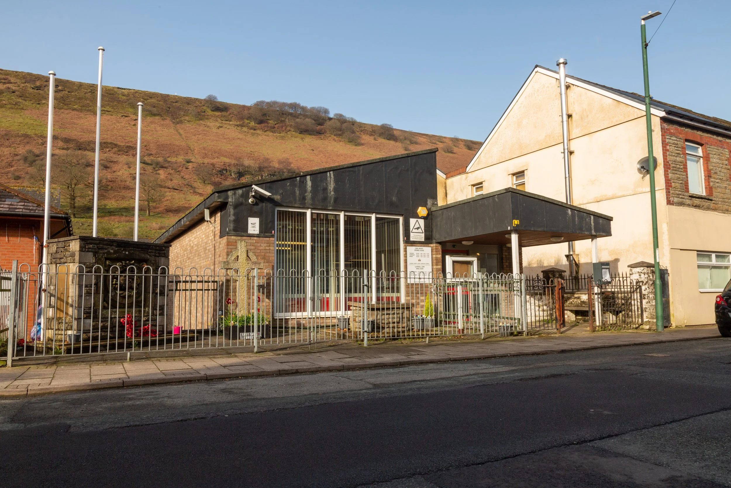 A small church building with a glass entrance, memorial cross, and roadside fence, against a hillside background on a clear day.