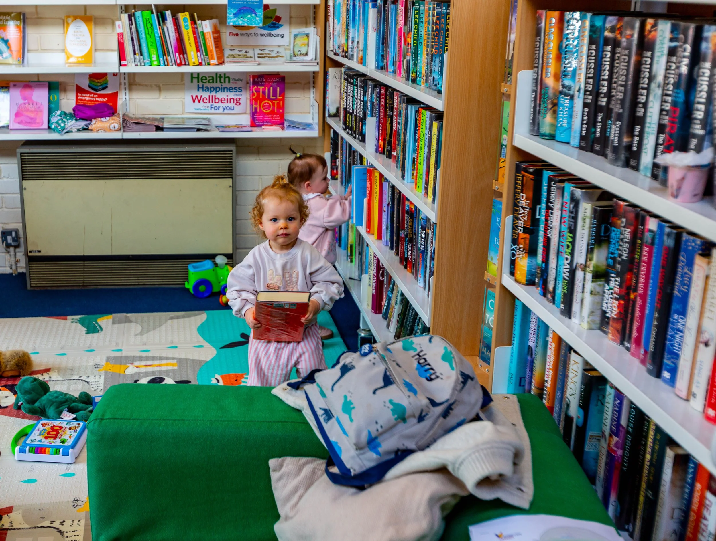 Two young children in a library, one holding a book and looking at the camera, while the other is on the bookshelves. The room has bookshelves filled with colorful books, a green table with a backpack and some clothes, and a decoupage rug with variou