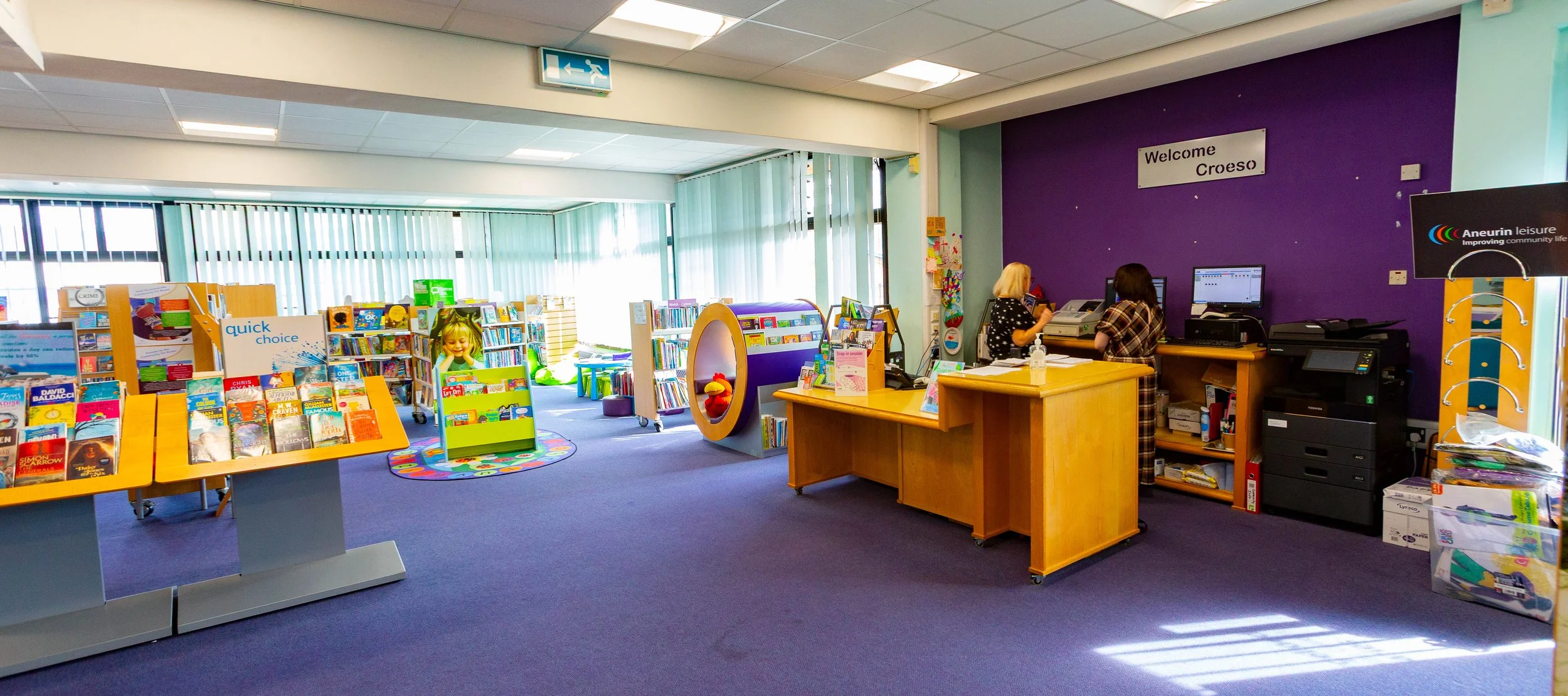 Interior of a community library with children's books and a play area. Front displays shelves of books and a circular book display. Background shows knitting area with two staff members at a desk, purple wall with a 'Welcome Croeso' sign, and an exit