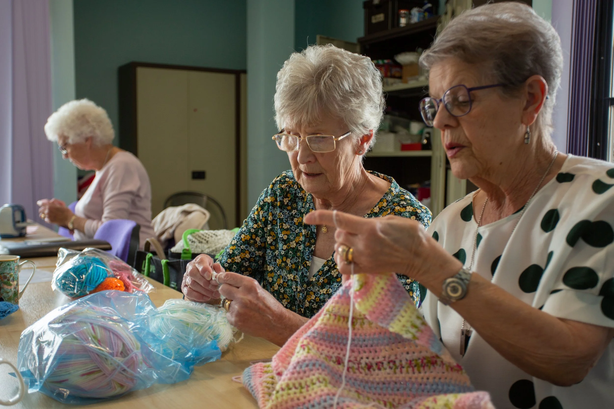 Three elderly women sitting at a table engaged in knitting and crocheting, with colorful yarn and knitting needles visible.