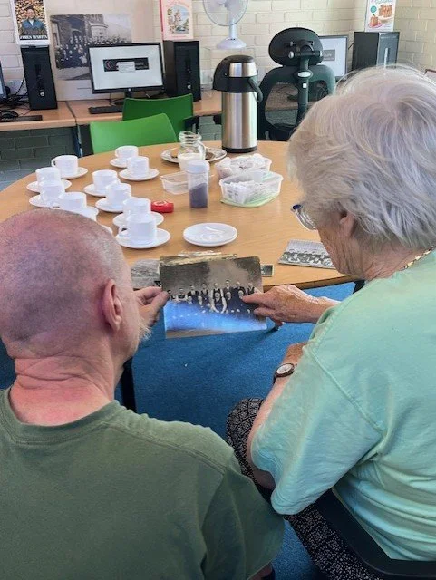 Two elderly people, a man and a woman, are sitting at a table looking at a photograph or a small picture. The table has several empty cups and a thermos on it. In the background, there are computer monitors, a fan, and various items.