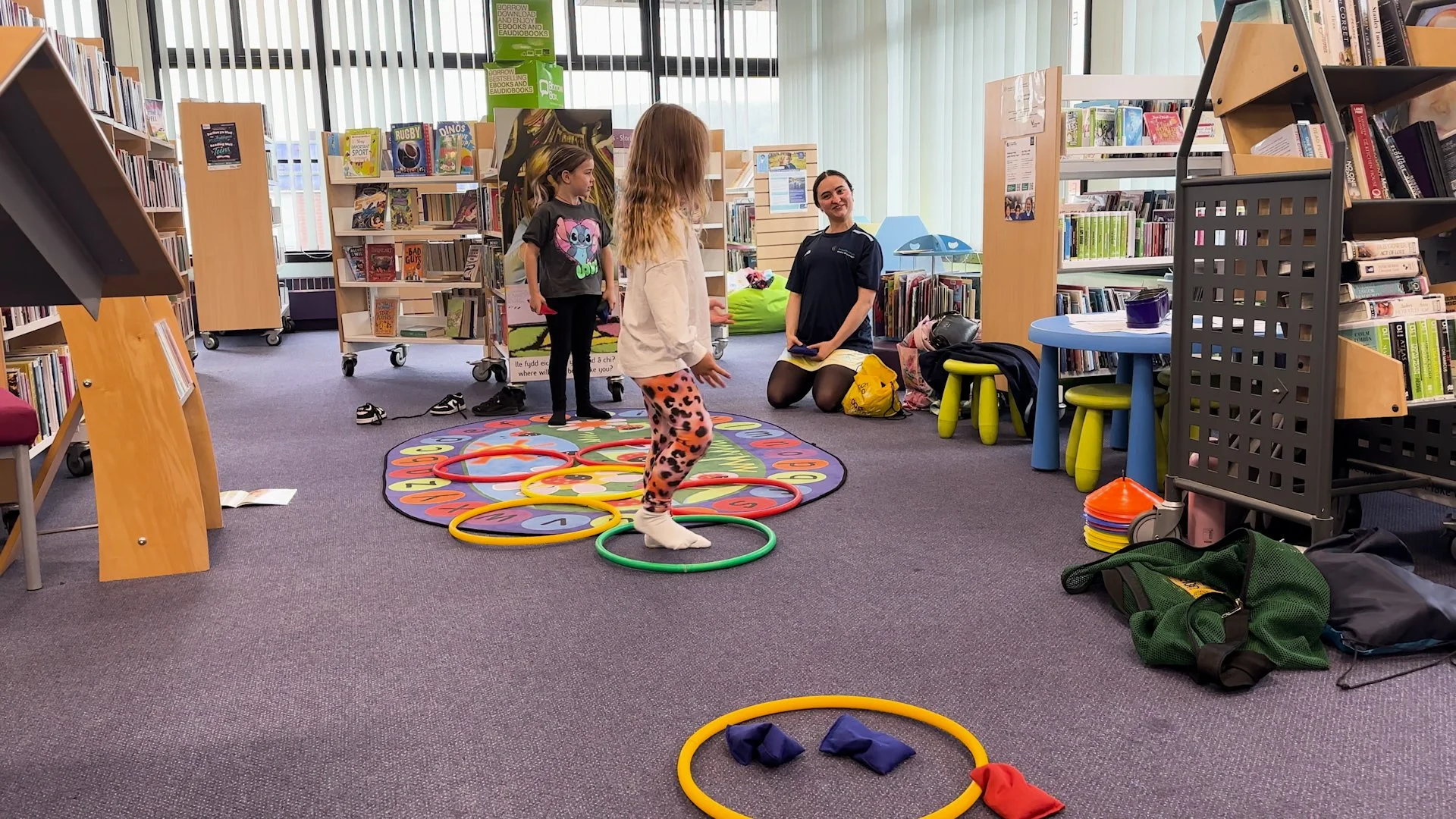 Children playing a ring toss game on a colorful carpet inside a library, with a woman supervising and bookshelves in the background.